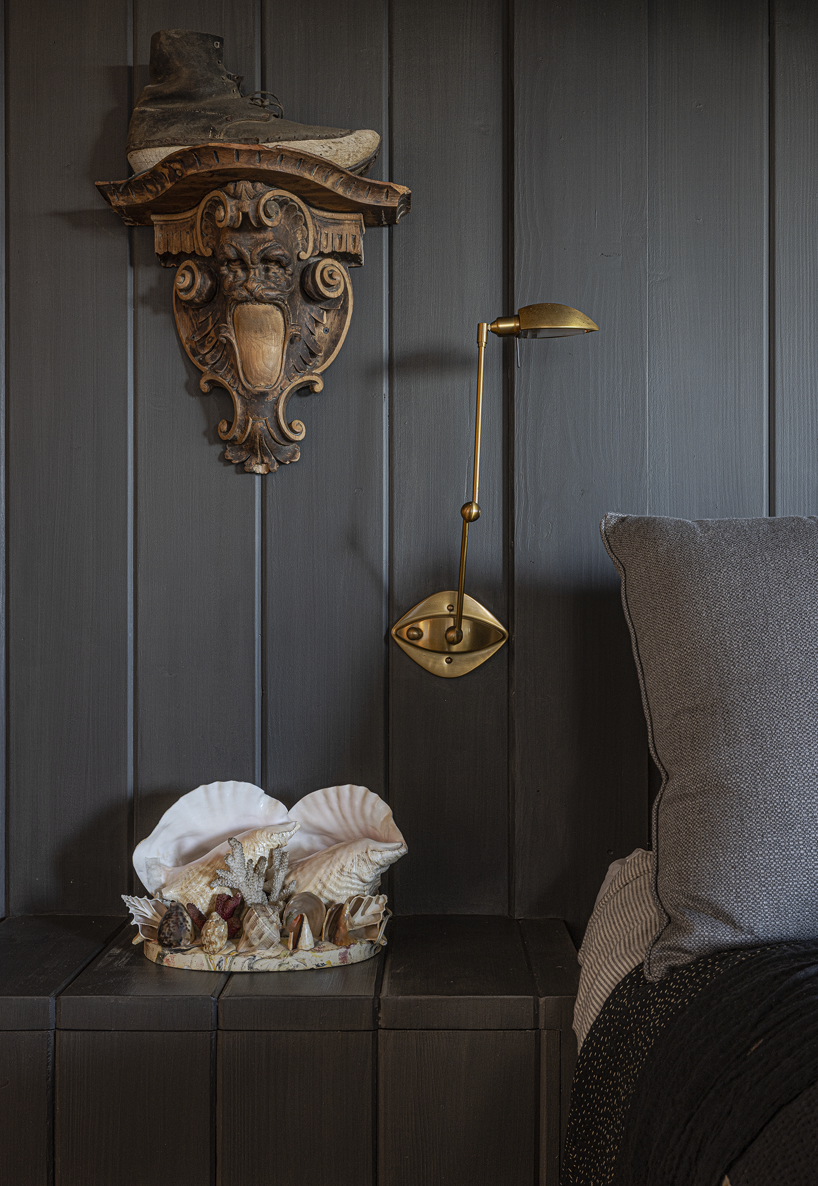 Decorative wall with a vintage shoe on a carved wooden plaque, a brass wall lamp, and a shell collection on a dark wooden nightstand next to a gray upholstered bed.