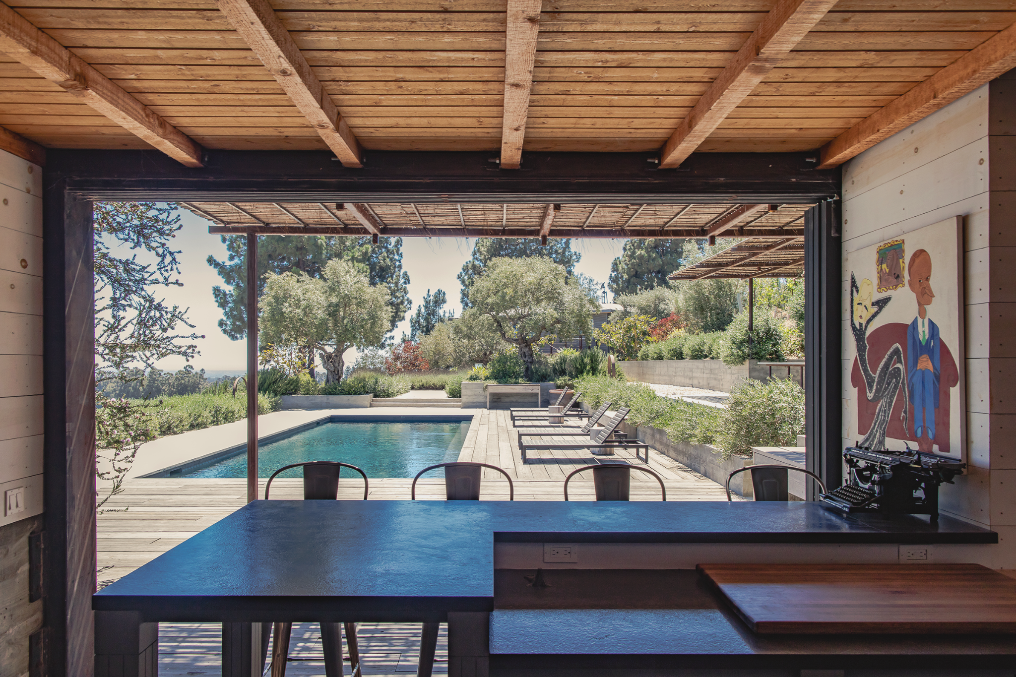 View from inside a house looking out onto a backyard with a swimming pool, lounge chairs, trees, and shrubs.