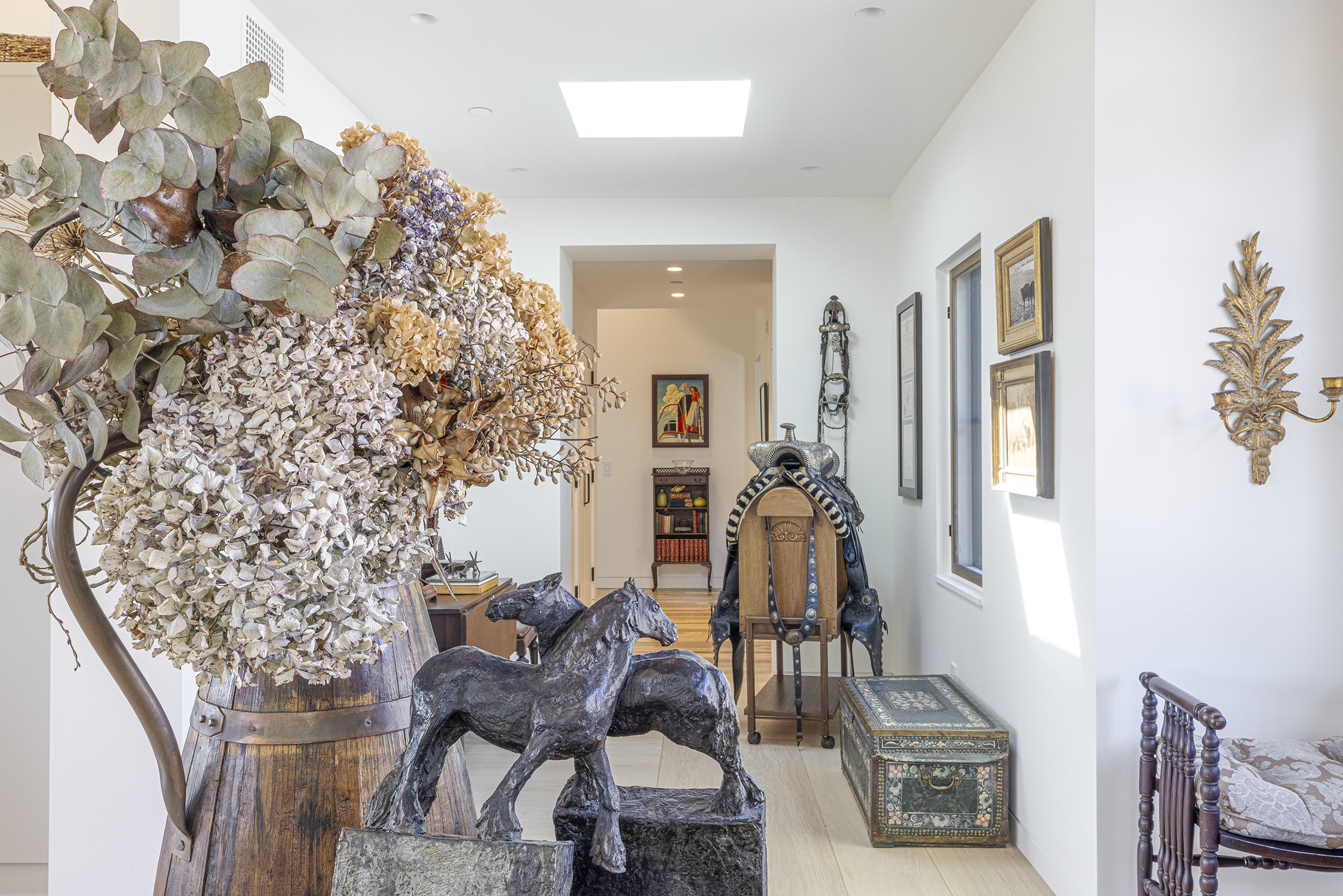 Interior view of a bright hallway or living space with white walls, wooden flooring, and various decorative items including dried flowers, sculptures, and framed artwork.