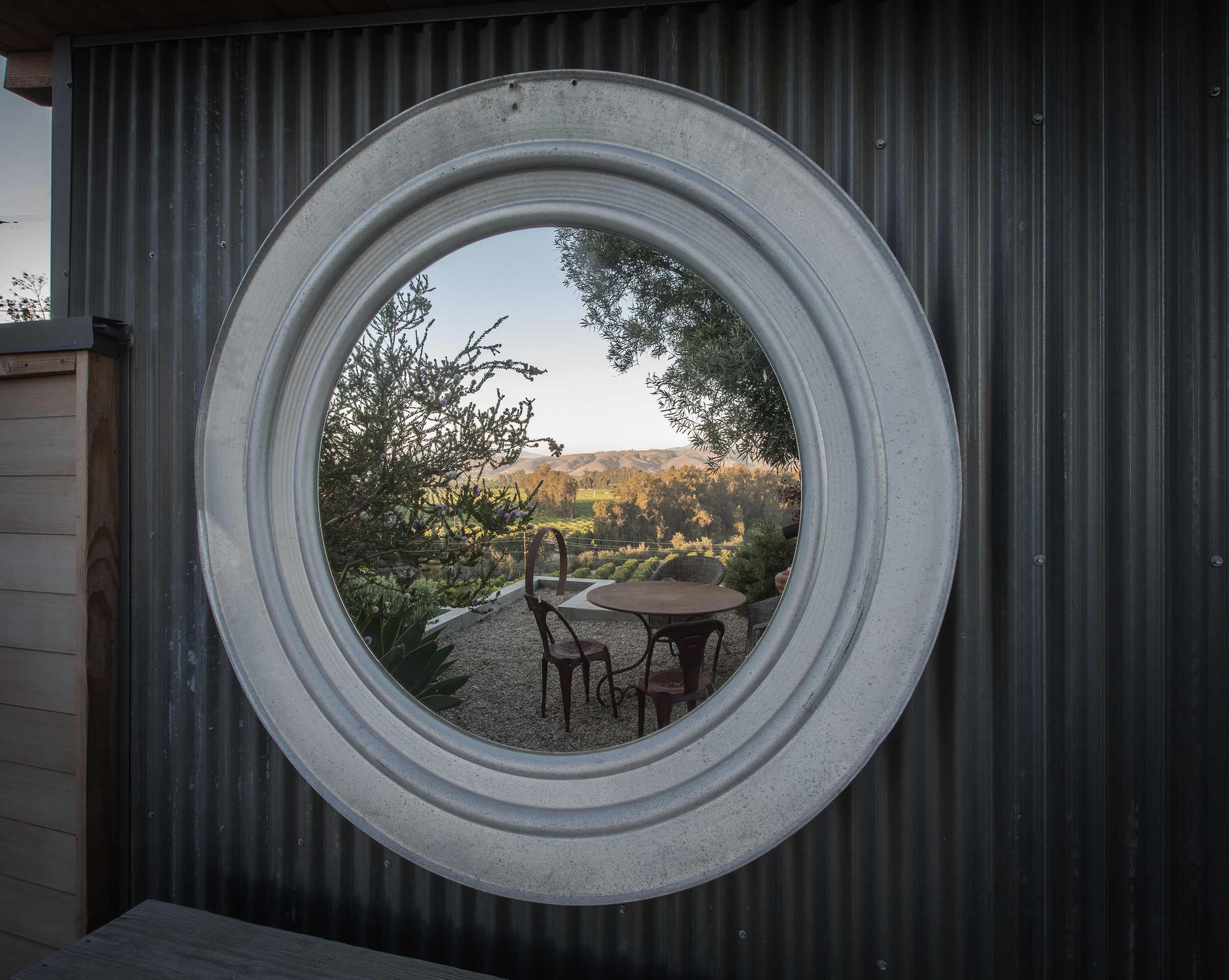 View of an outdoor patio with a round table and chairs, seen through a circular window in a corrugated metal wall, with trees and rolling hills in the background.