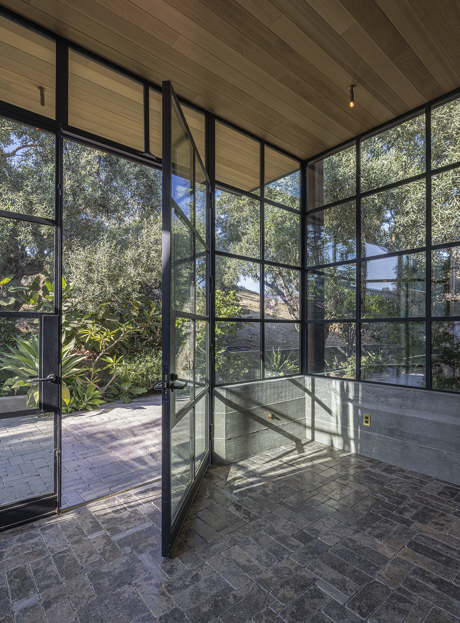 Interior view of a modern sunroom with floor-to-ceiling glass windows, black metal framing, a brick floor, and a wooden ceiling, overlooking lush greenery outside.