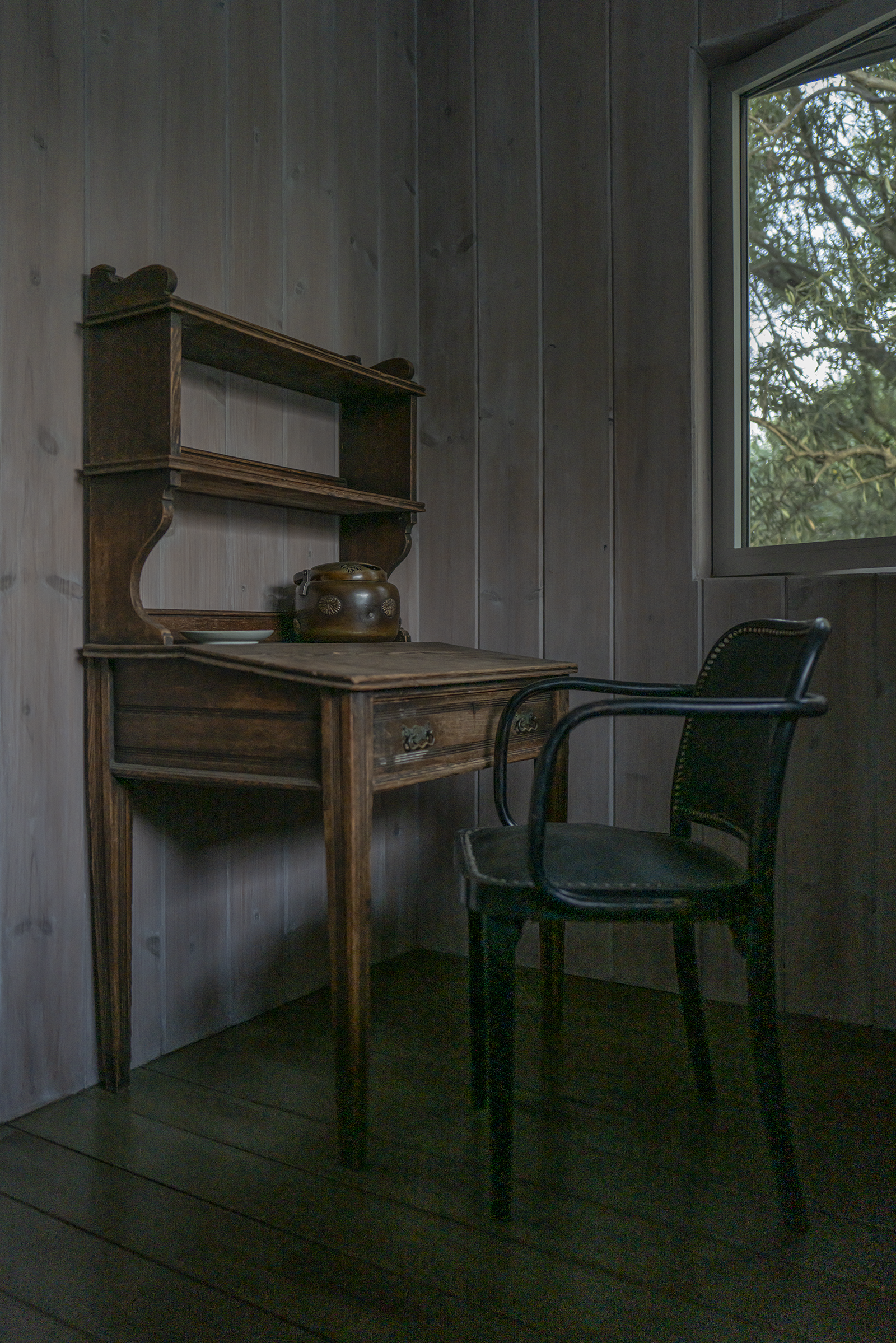 A corner of a room with wood-paneled walls, a wooden desk, a black chair, a window showing trees outside, and a decorative jar on the desk.