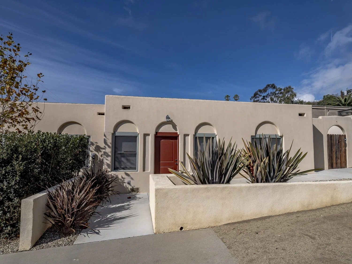 A beige stucco building with arched windows and a red door, surrounded by desert plants and bushes, under a blue sky with clouds.