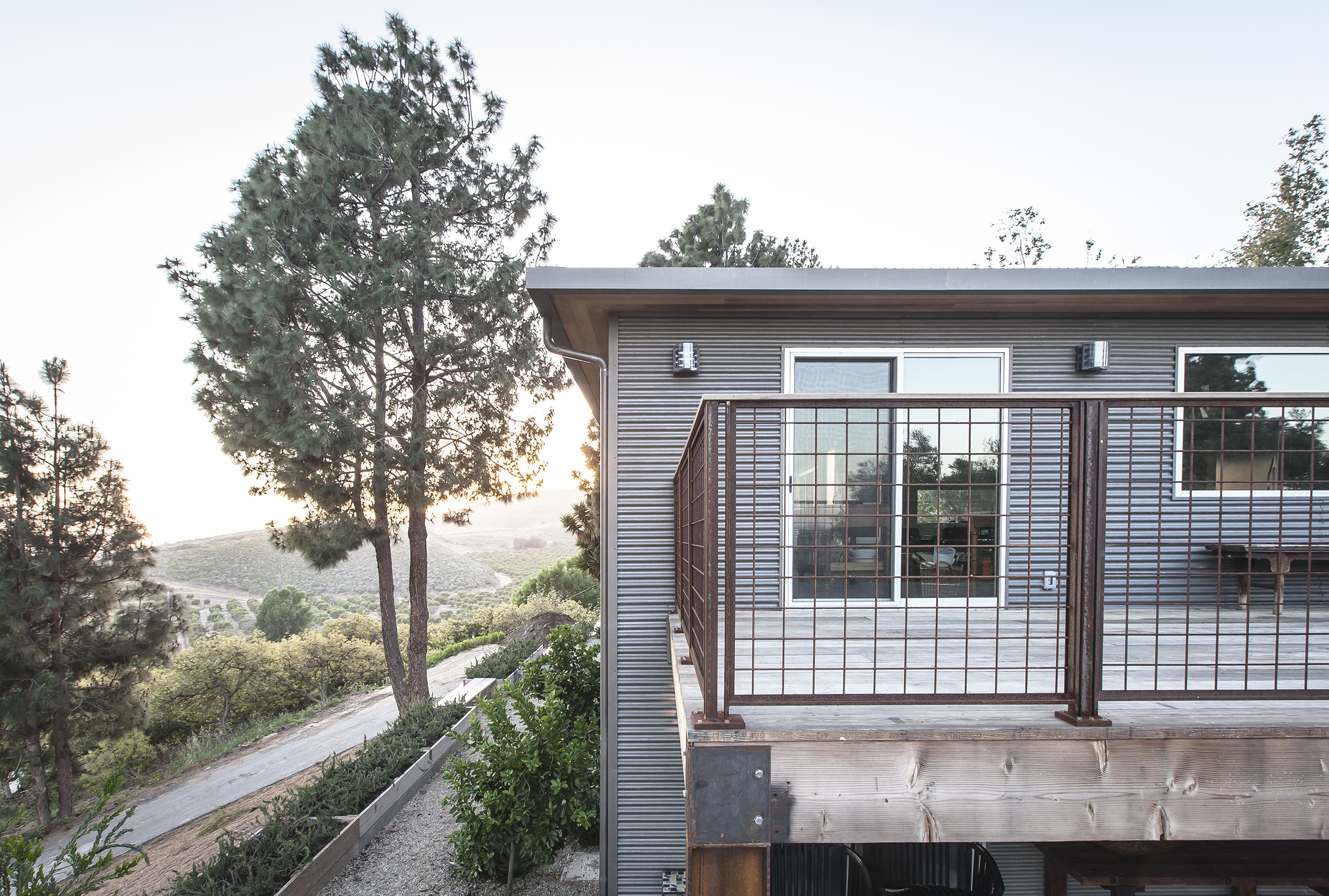 Exterior view of a modern house with a balcony, metal railing, large window and sliding glass door, surrounded by trees and a hillside at sunset.