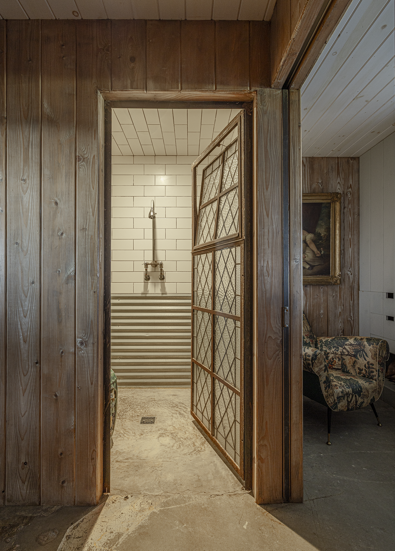View into a vintage-style shower with white subway tiles, a metal showerhead, and a striped radiator, separated by a rustic, divided glass door, surrounded by wooden paneling and a glimpse of a patterned armchair and framed painting in an adjacent ro