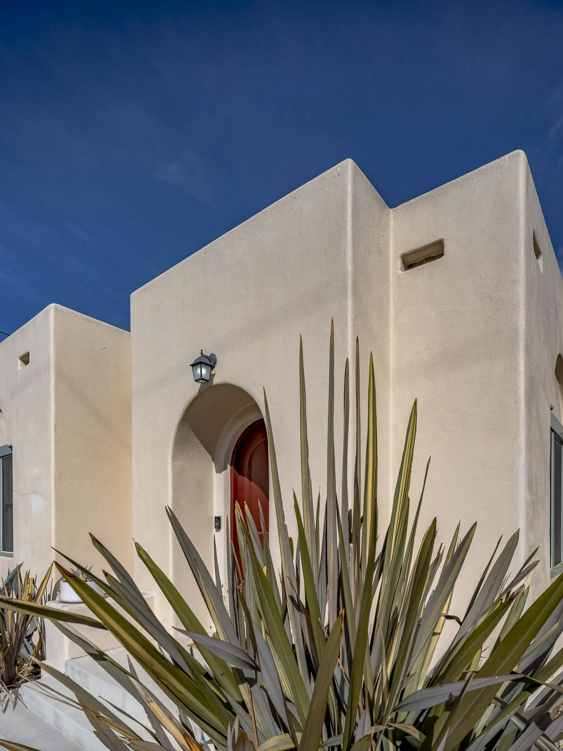 Modern white stucco house with arched doorway and red door, large spiky plant in front, clear blue sky in background.