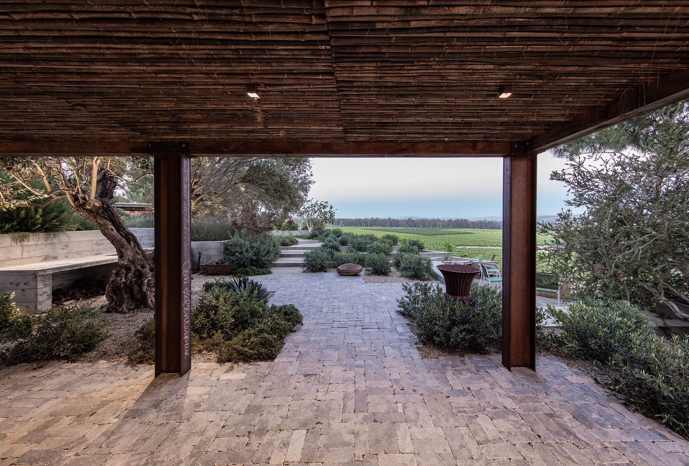 View from under a wooden overhang onto a patio with brick paving, green shrubs, trees, and a landscape of fields in the distance.