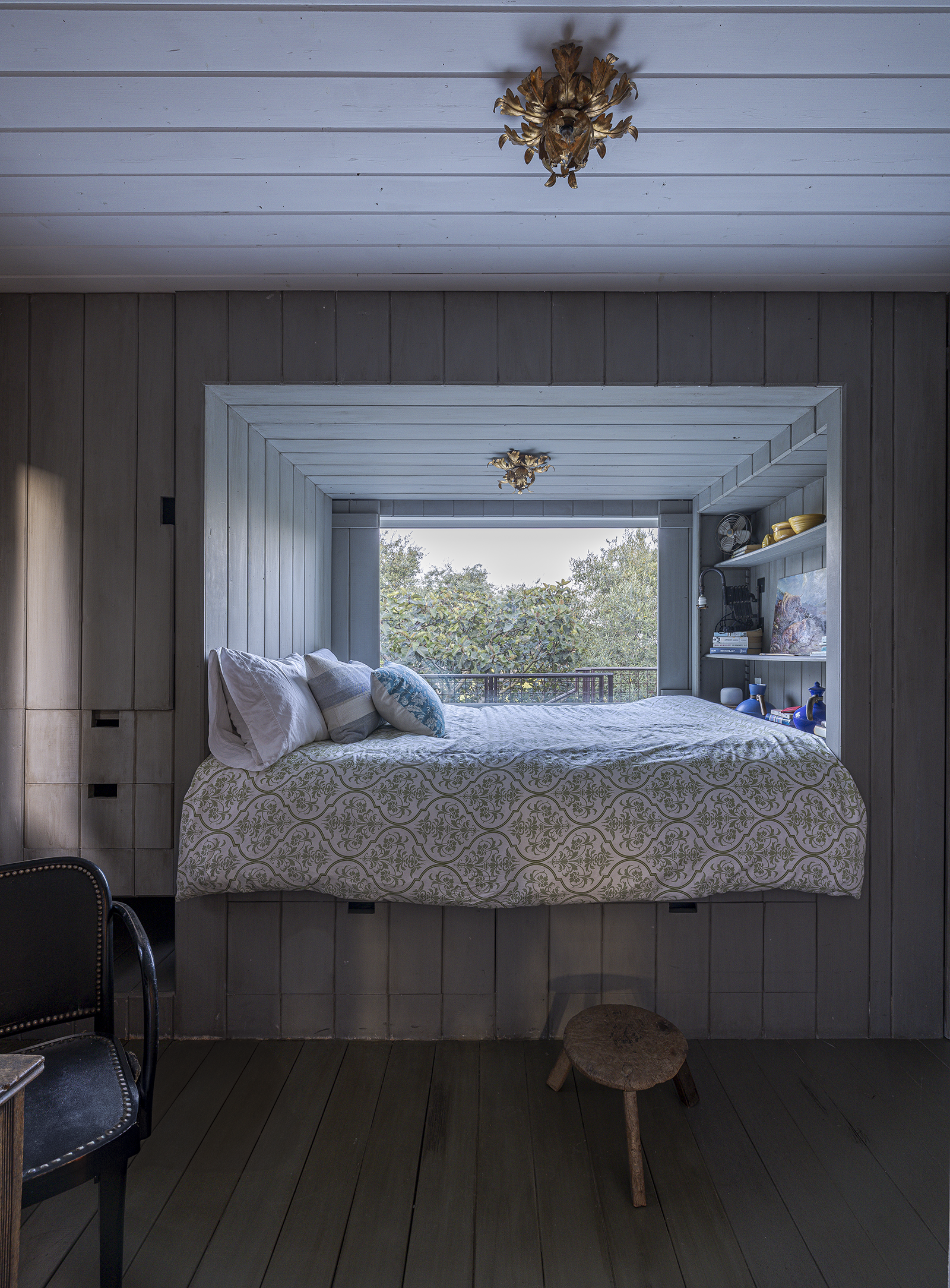 A bedroom with a window seat bed covered in white and gold patterned bedding, pillows, and a view of trees outside. The room has wood-paneled walls and a ceiling with decorative brass fixtures.