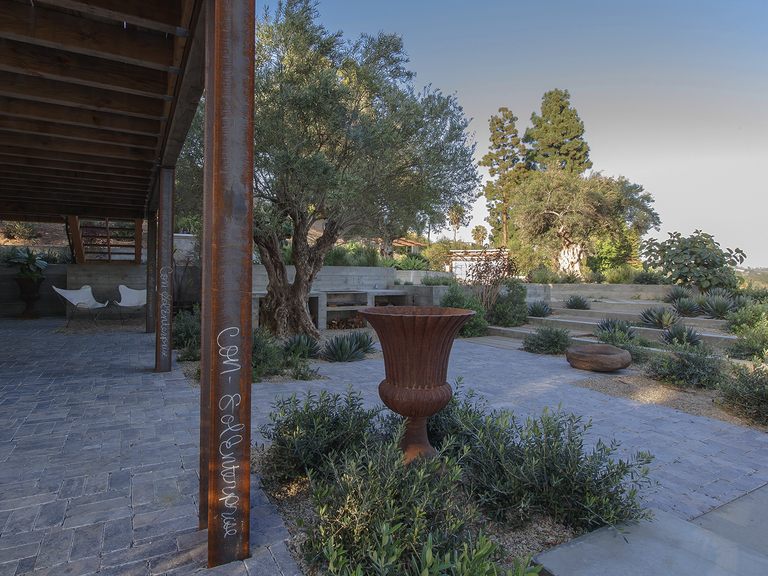 An outdoor landscaped patio area with stone paving, a rust-colored metal planter, trees, shrubs, and a seating area under a wooden awning.