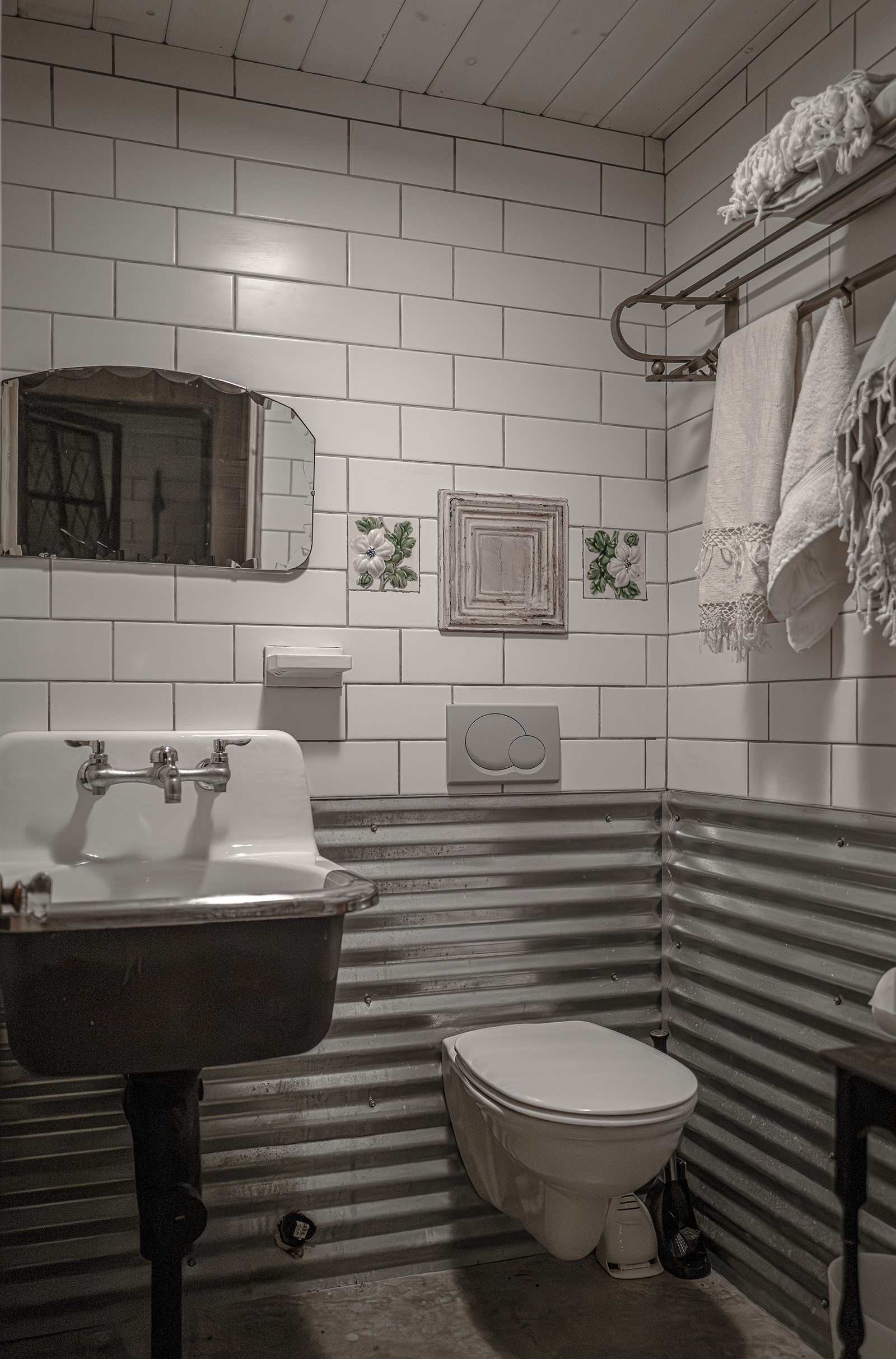 A small bathroom with white subway tiles, a black and white sink, a toilet, a mirror, and towels hanging on a metal rod. The lower part of the walls is made of corrugated metal.