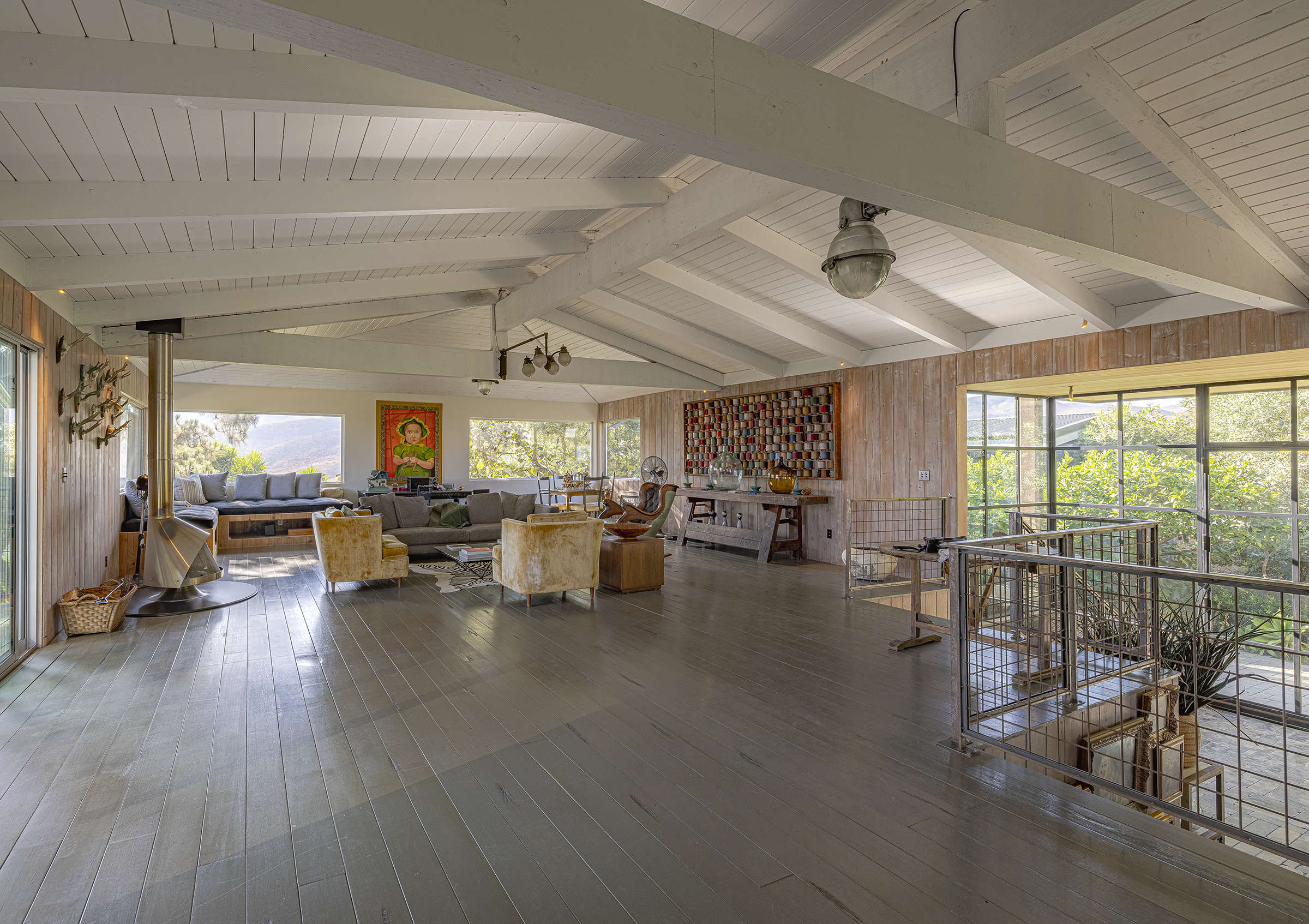 Spacious living room with wooden flooring, large windows with greenery outside, a cozy seating area with couches and armchairs, decorative artwork on the walls, and a staircase railing near the entrance.