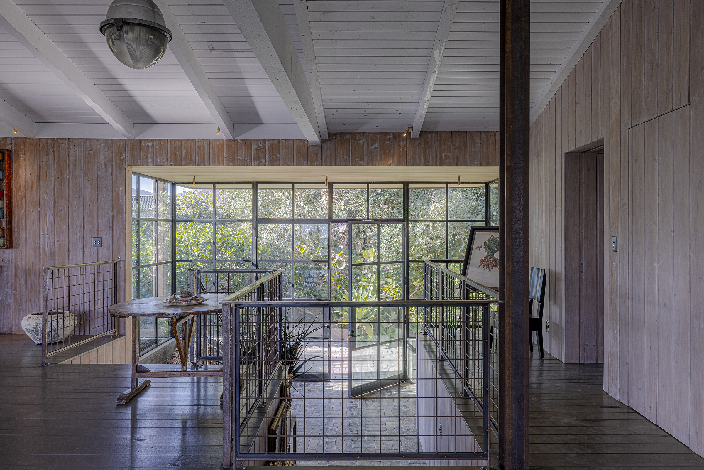 Interior view of a room with wood-paneled walls, white painted ceiling with exposed beams, a large window with a garden view, a metal railing, a small table, a framed artwork, and various decorative items.