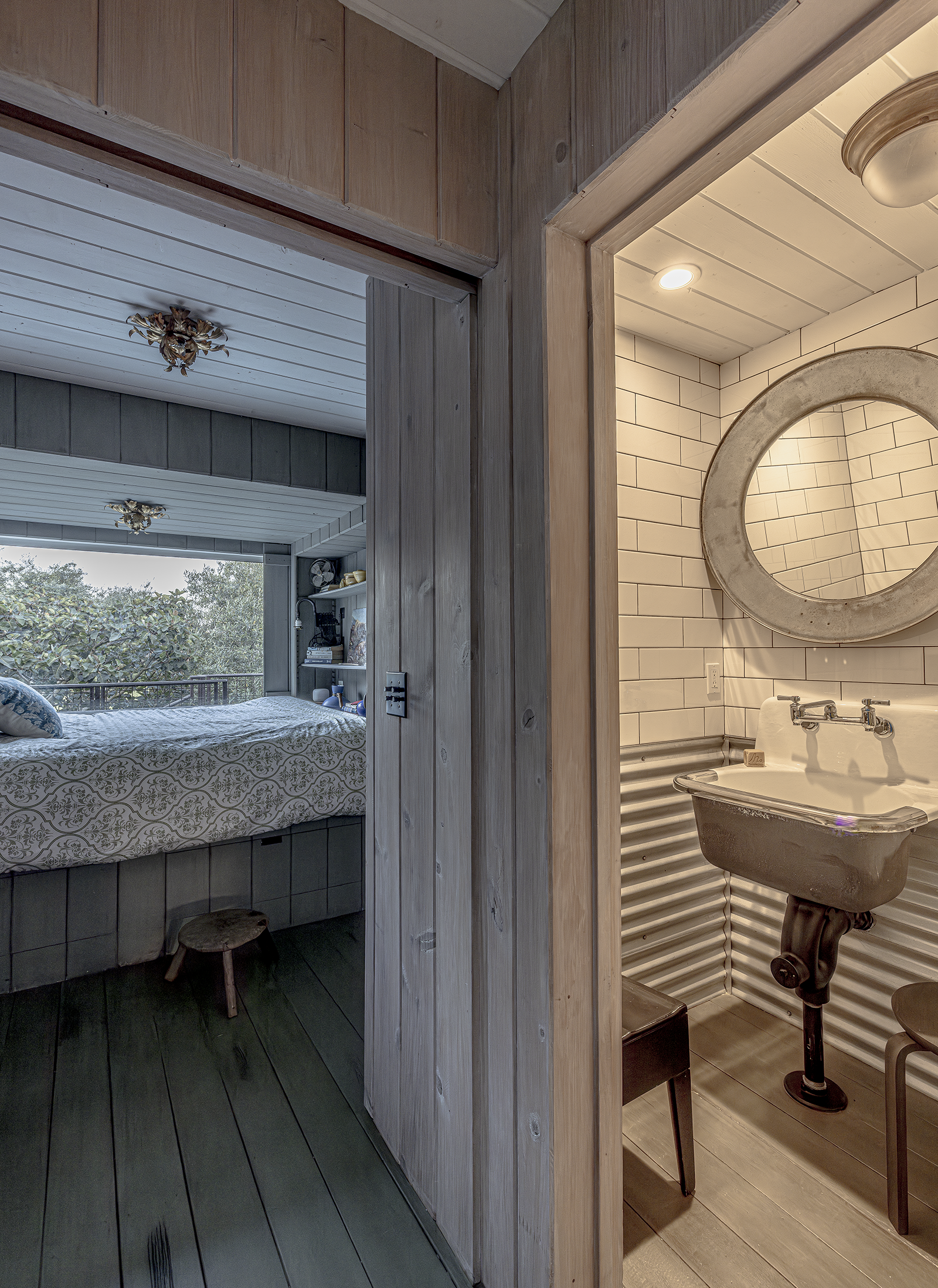 Interior of a cozy bedroom with gray wood paneling, a large window with a view of trees, and a bed with a patterned quilt. Adjacent to the bedroom is a small rustic bathroom with white subway tiles, a round mirror, a vintage sink, and two chairs.