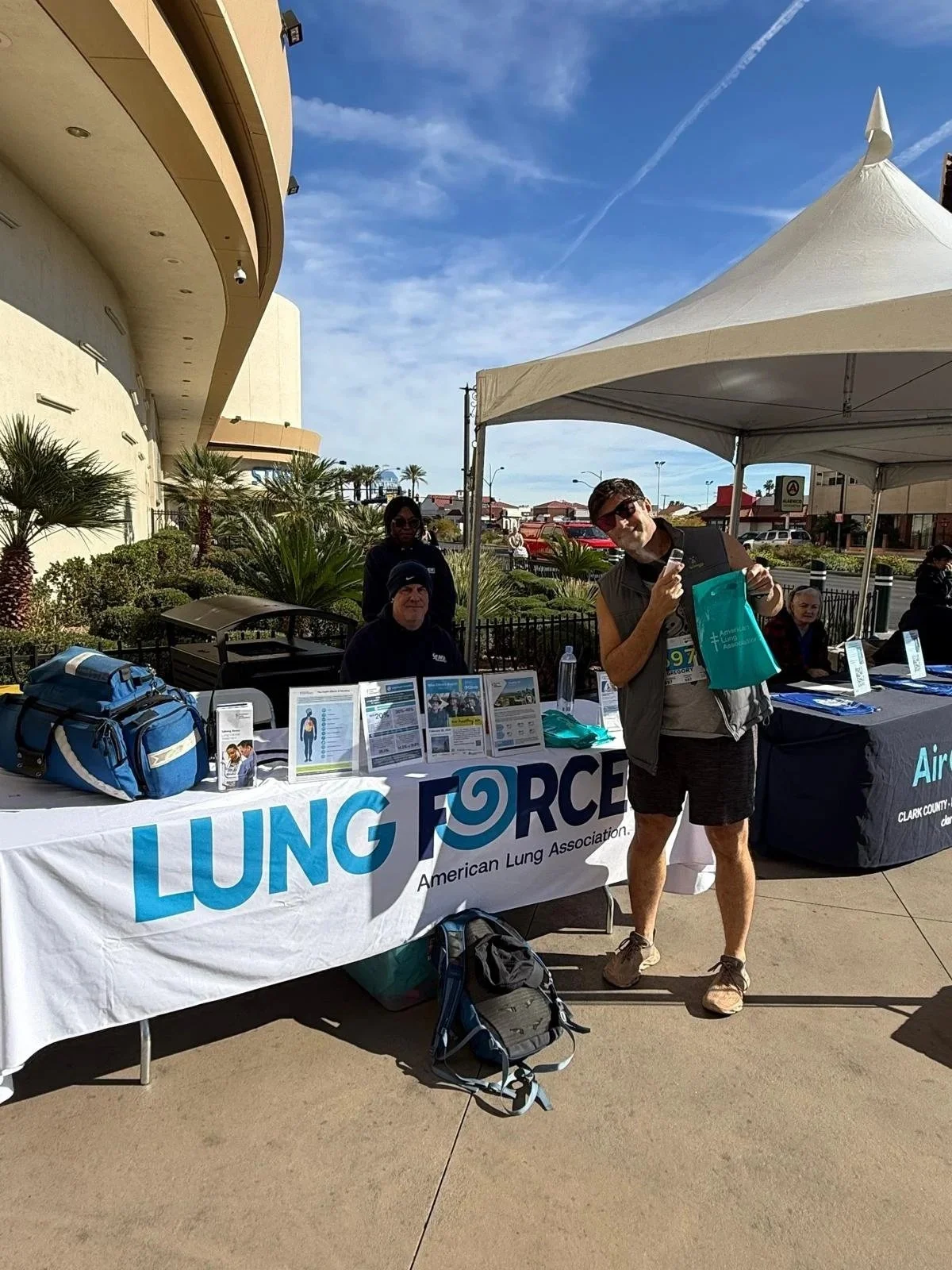 An outdoor event with informational tables and a tent for the American Lung Association called 'LUNG FORCE.' People are sitting and standing, some holding informational materials, in a sunny area with palm trees and buildings in the background.
