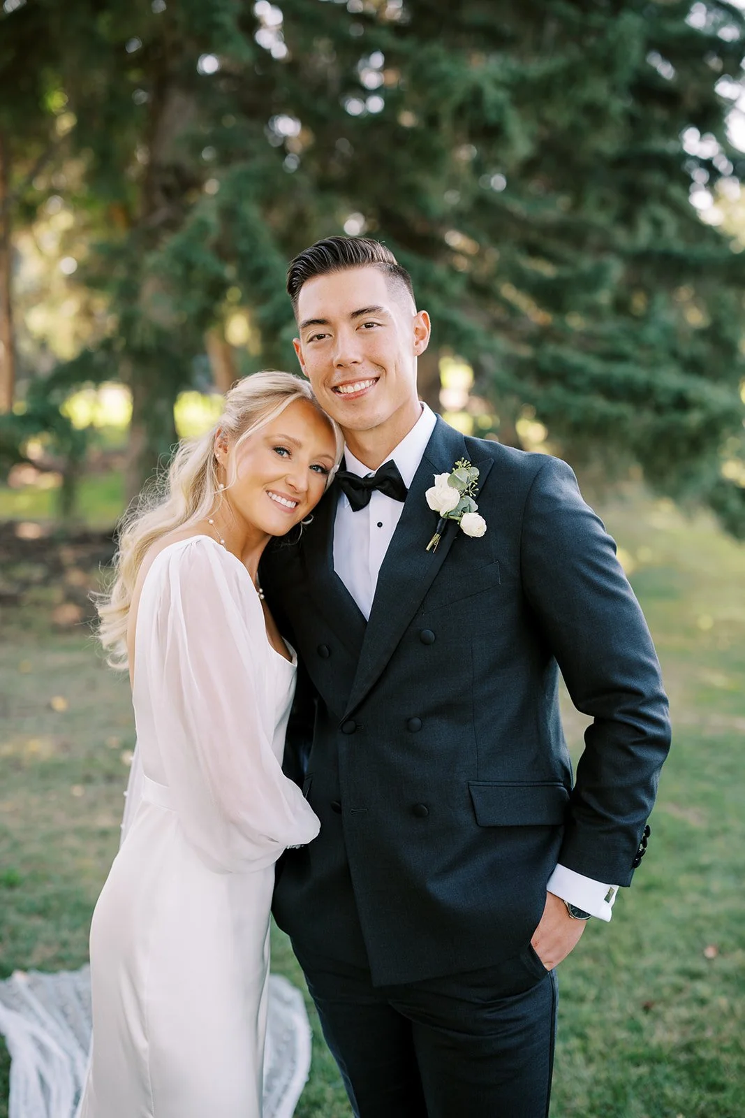 A smiling couple, possibly bride and groom, with the woman in a white dress and the man in a black suit and bow tie, standing outdoors with trees in the background.