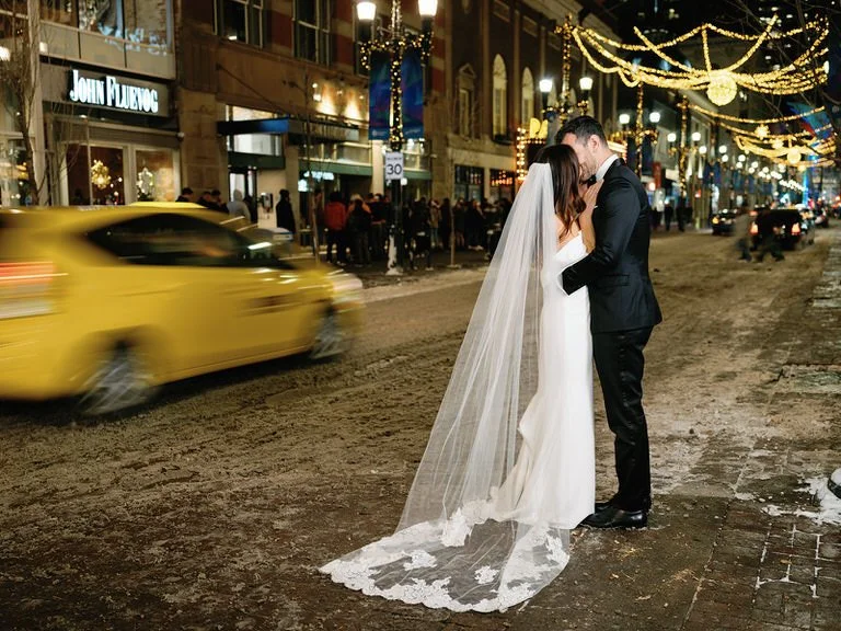 Bride and groom kissing on a city street at night with holiday lights and a yellow taxi passing by.