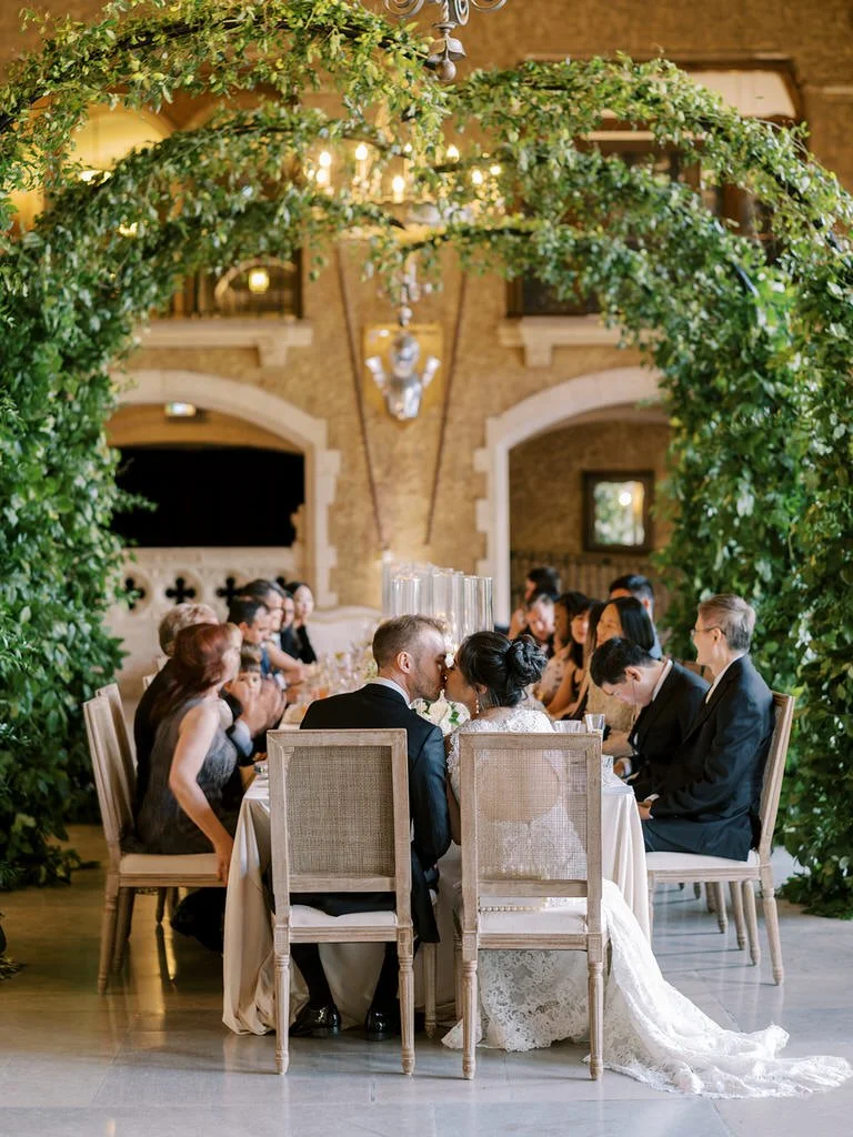 Wedding reception with newlyweds kissing, seated at a table under a lush greenery arch, surrounded by guests in a decorated venue.