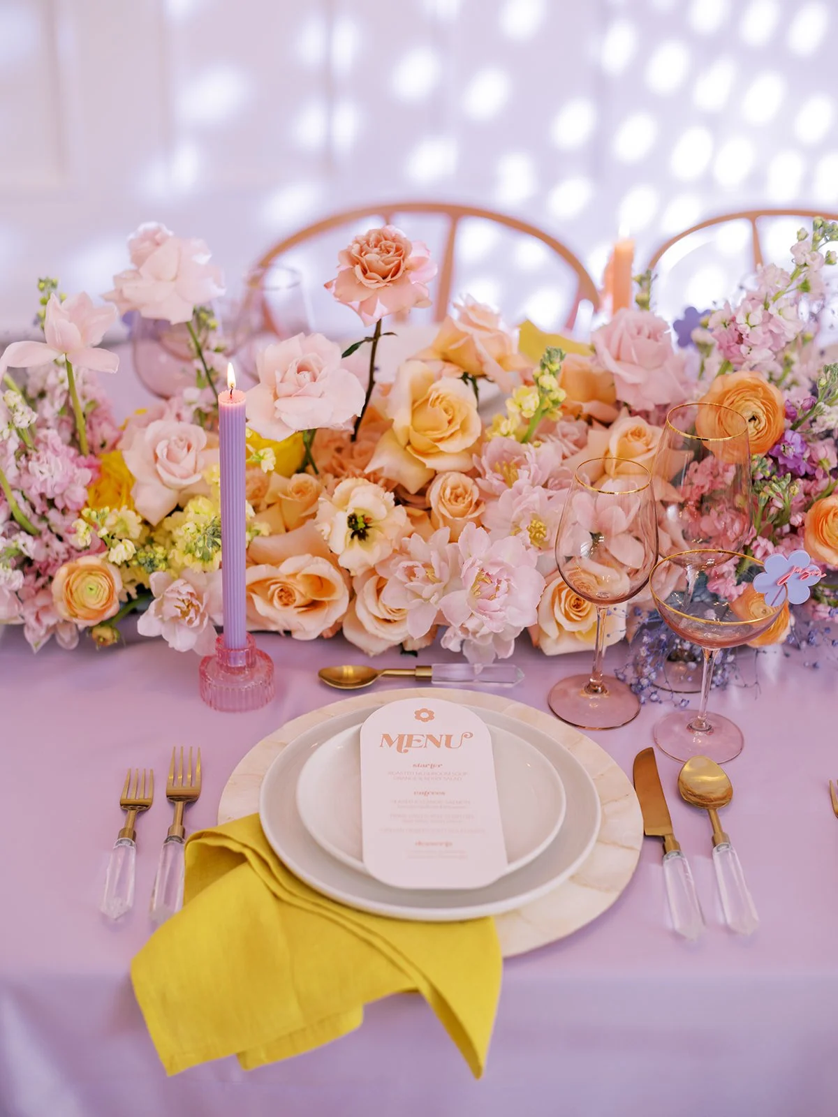 Elegant table setting with colorful floral arrangement, purple candle, yellow napkin, and gold cutlery on a lavender tablecloth. Includes a plate with a menu card.