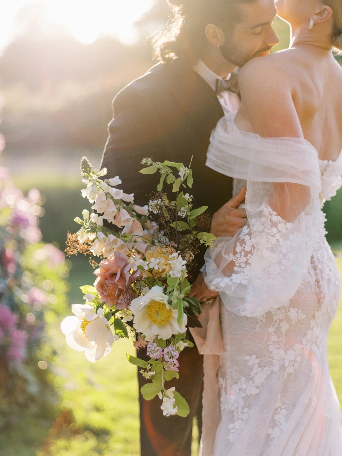 Bride and groom embracing outdoors, holding a bouquet of white and pink flowers, with sunlight in the background.