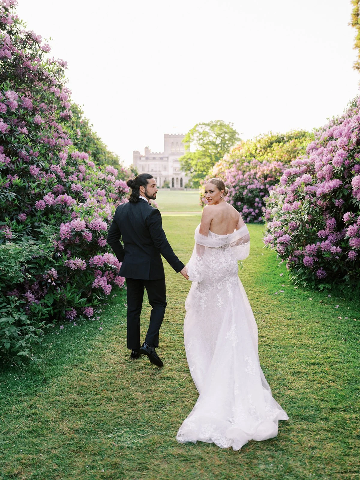 A bride and groom walking hand in hand down a garden path lined with purple flowering bushes, with a castle visible in the background.