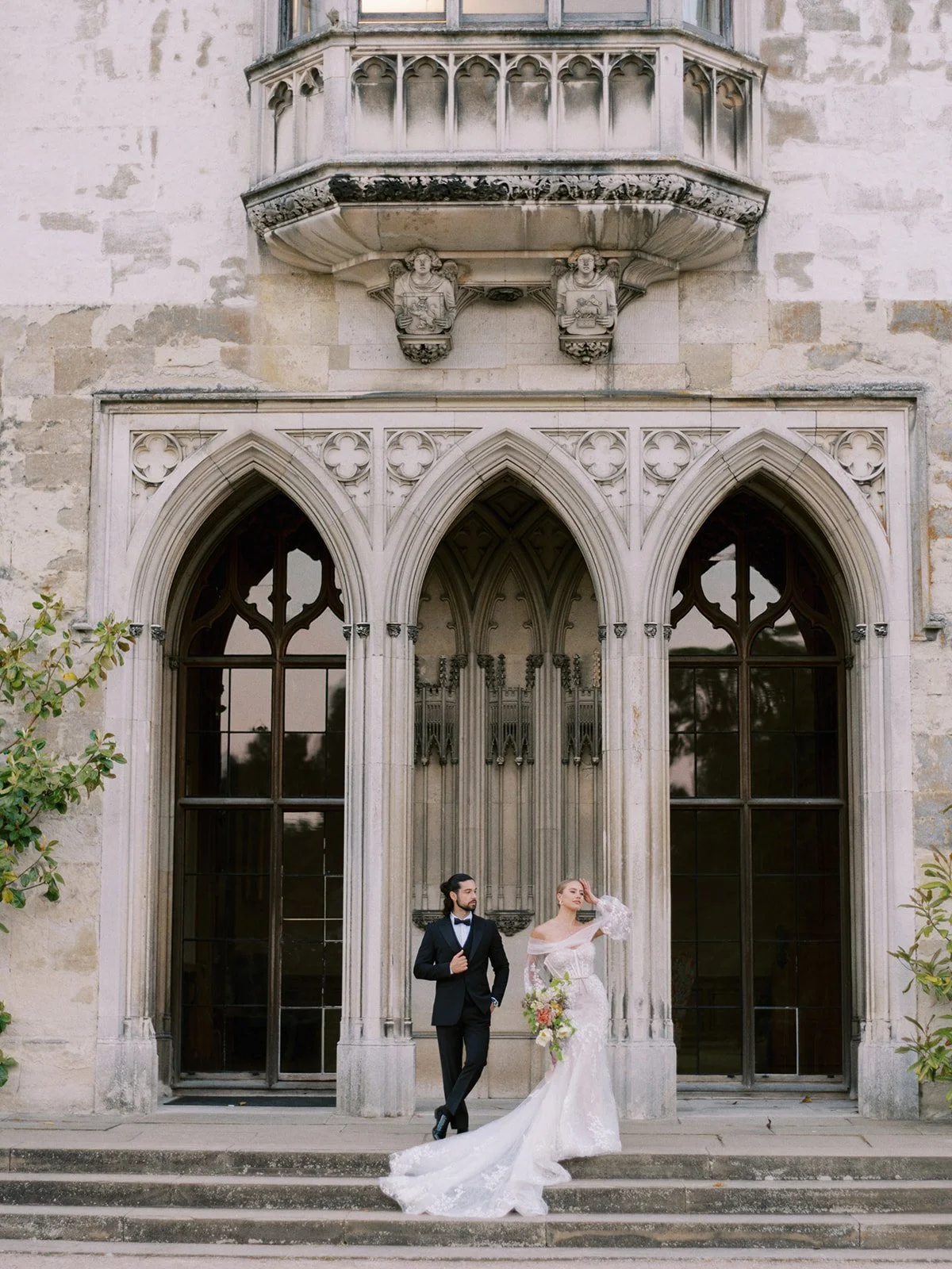 Bride and groom in front of a historic building with large arched windows and stone carvings.