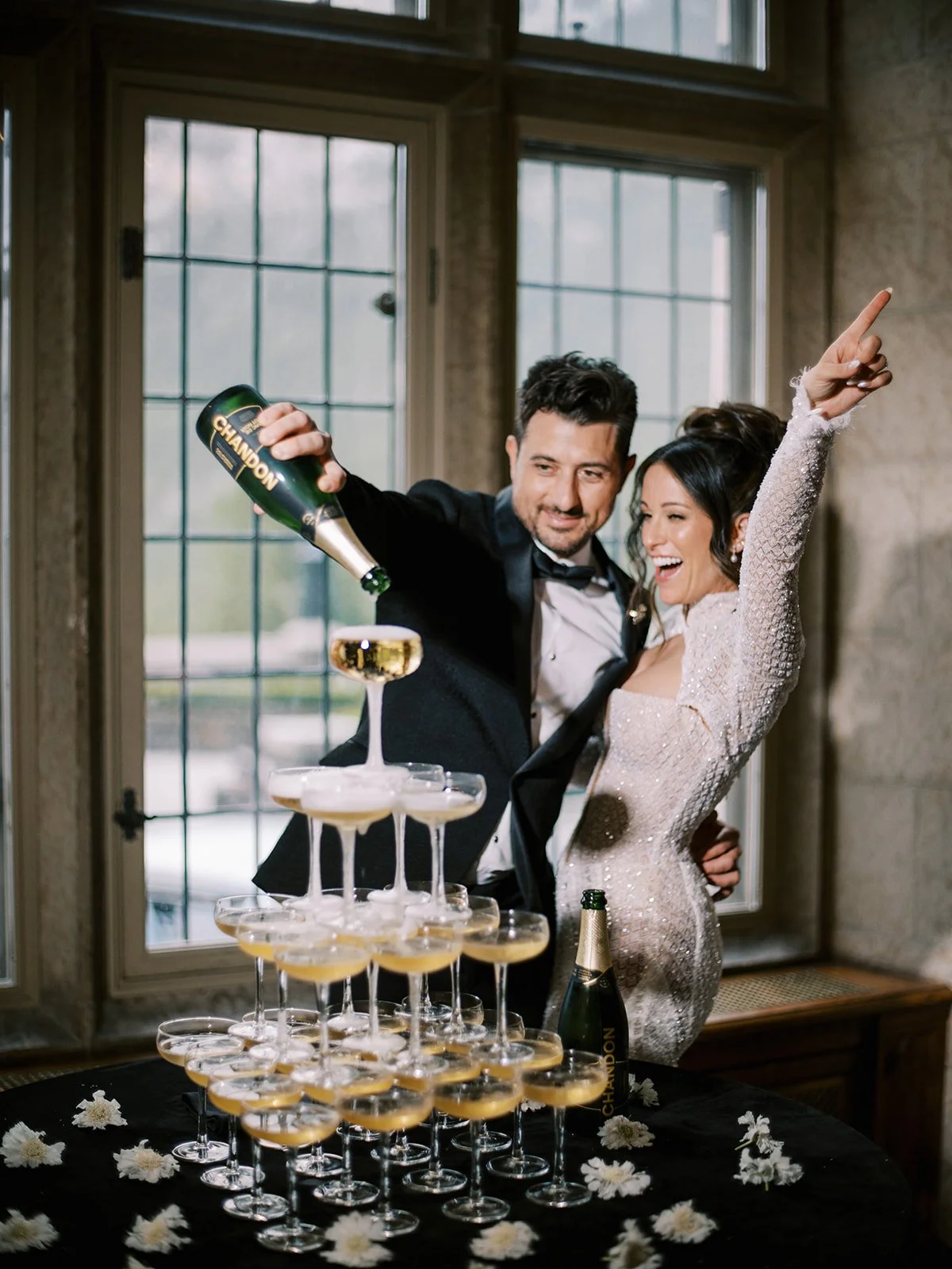A couple celebrating by pouring champagne into a champagne tower, dressed formally. The woman is wearing a sparkly dress and the man is in a suit with a bow tie. The table is decorated with flowers and a bottle of Chandon champagne is visible.