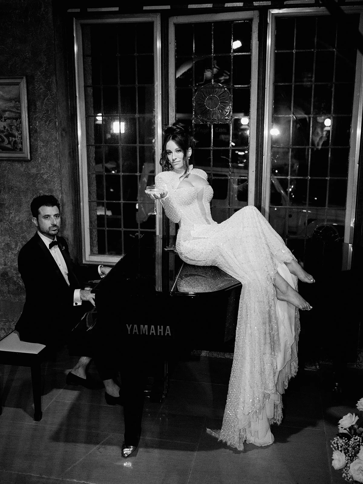 A woman in a long, elegant dress poses with a cocktail on a Yamaha grand piano, while a man in a tuxedo plays the piano. The setting is a stylish room with large windows and dim lighting.