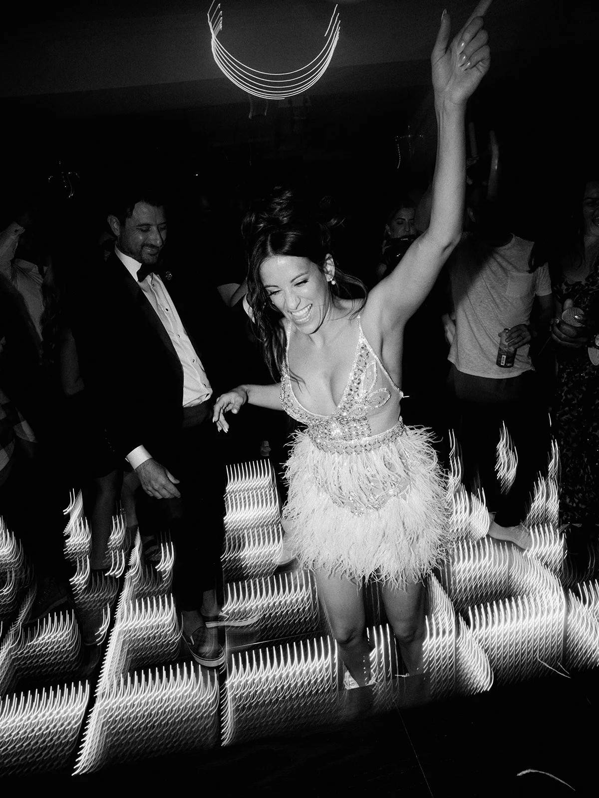 Black and white photo of a lively dance scene with a woman in a feathered dress joyfully dancing and pointing upwards, surrounded by people and motion blur lights.