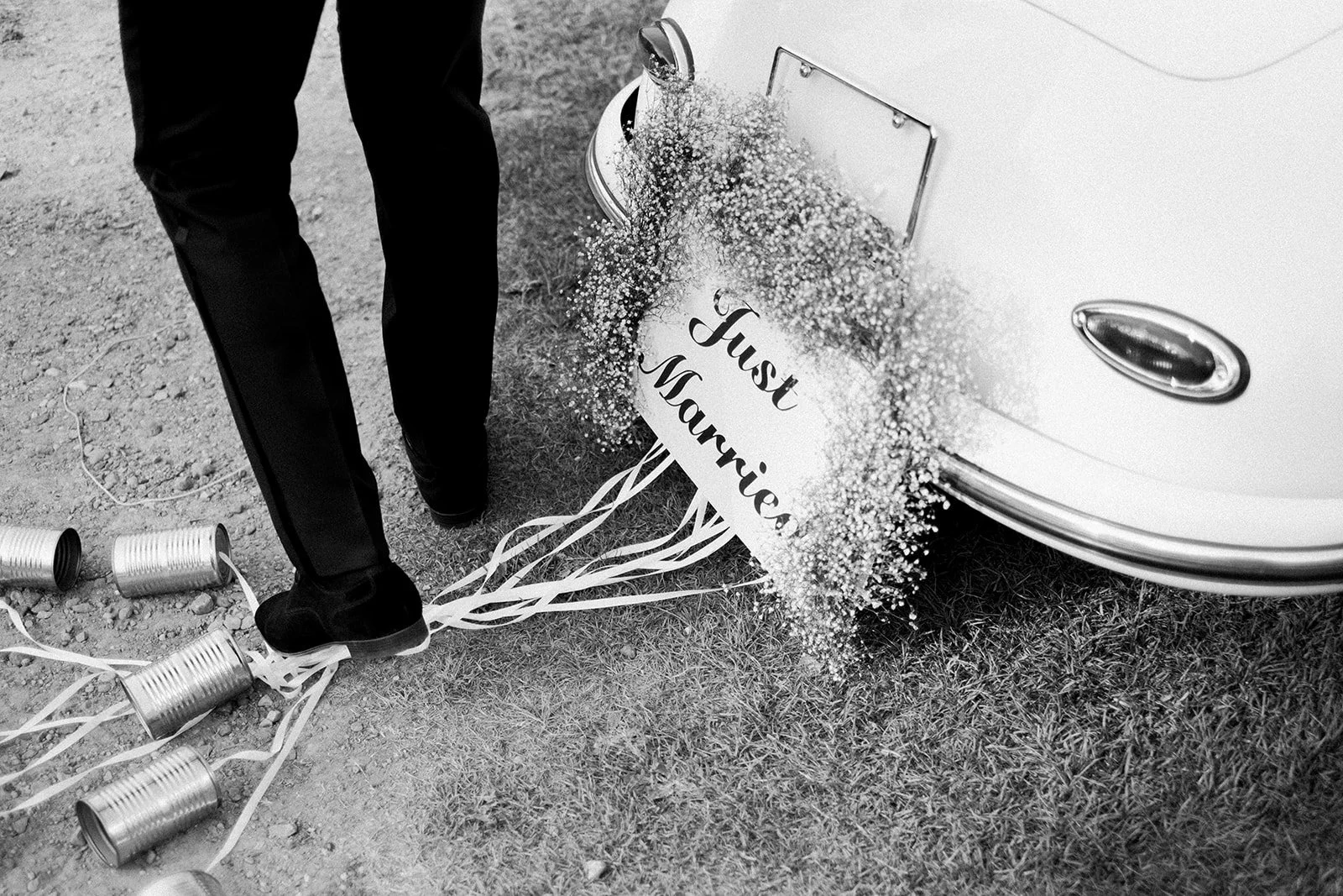 Black and white photo of a "Just Married" sign on the back of a car, with tin cans tied to it by ribbons, and a person stepping nearby on grass and dirt.