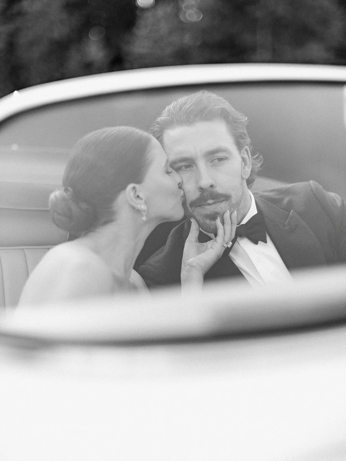 Black and white image of a woman kissing a man's cheek inside a car, both dressed formally.