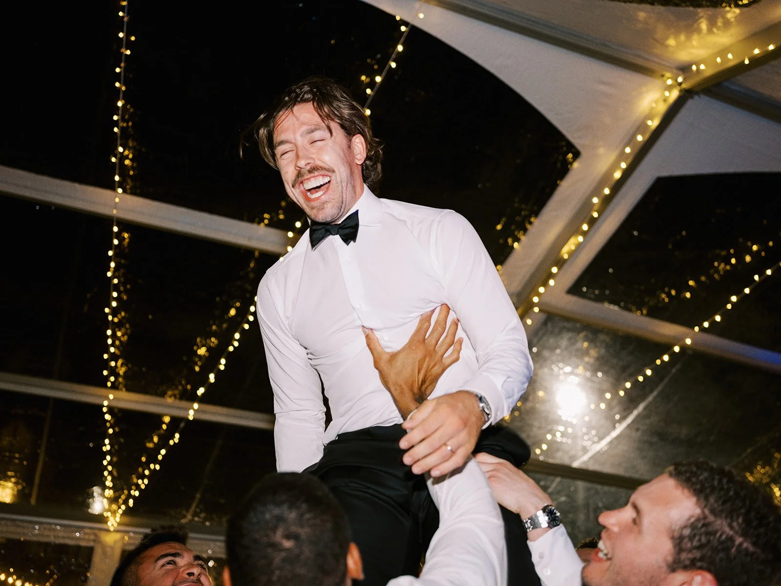 A joyful man in a white shirt and bow tie being lifted by others, surrounded by festive string lights.