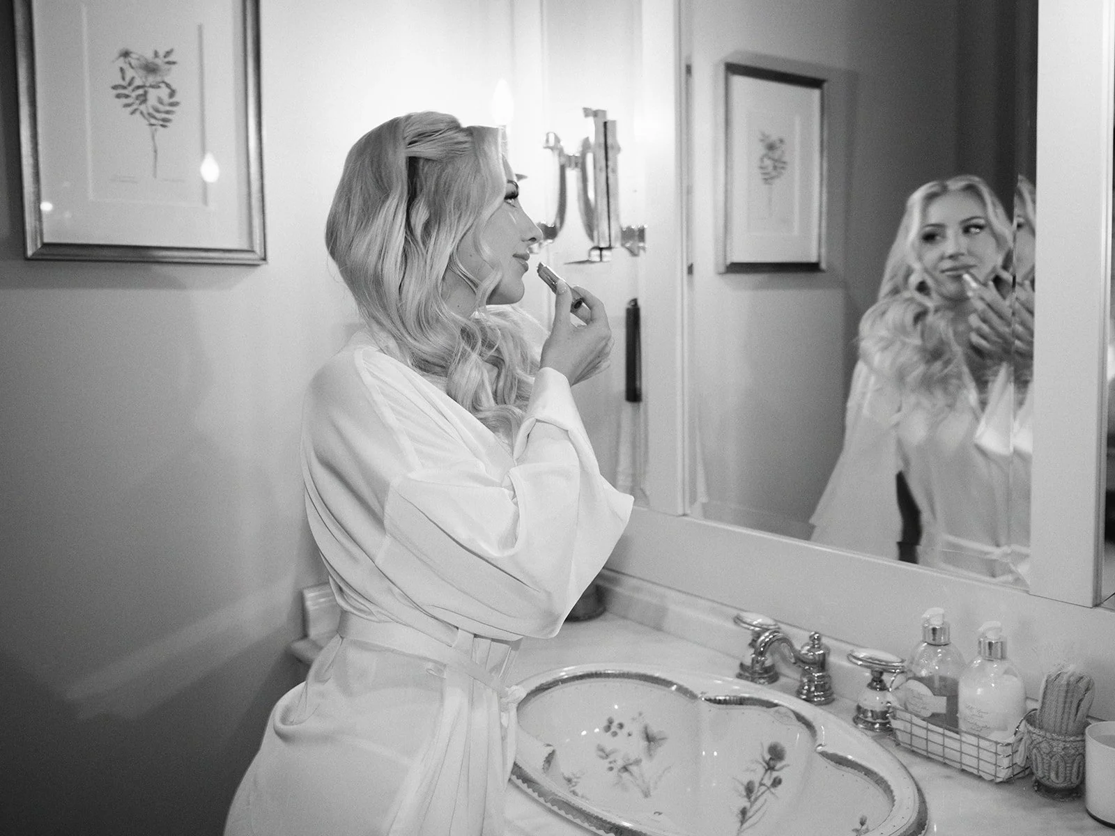 Black and white image of a woman in a robe applying lipstick in front of a bathroom mirror.