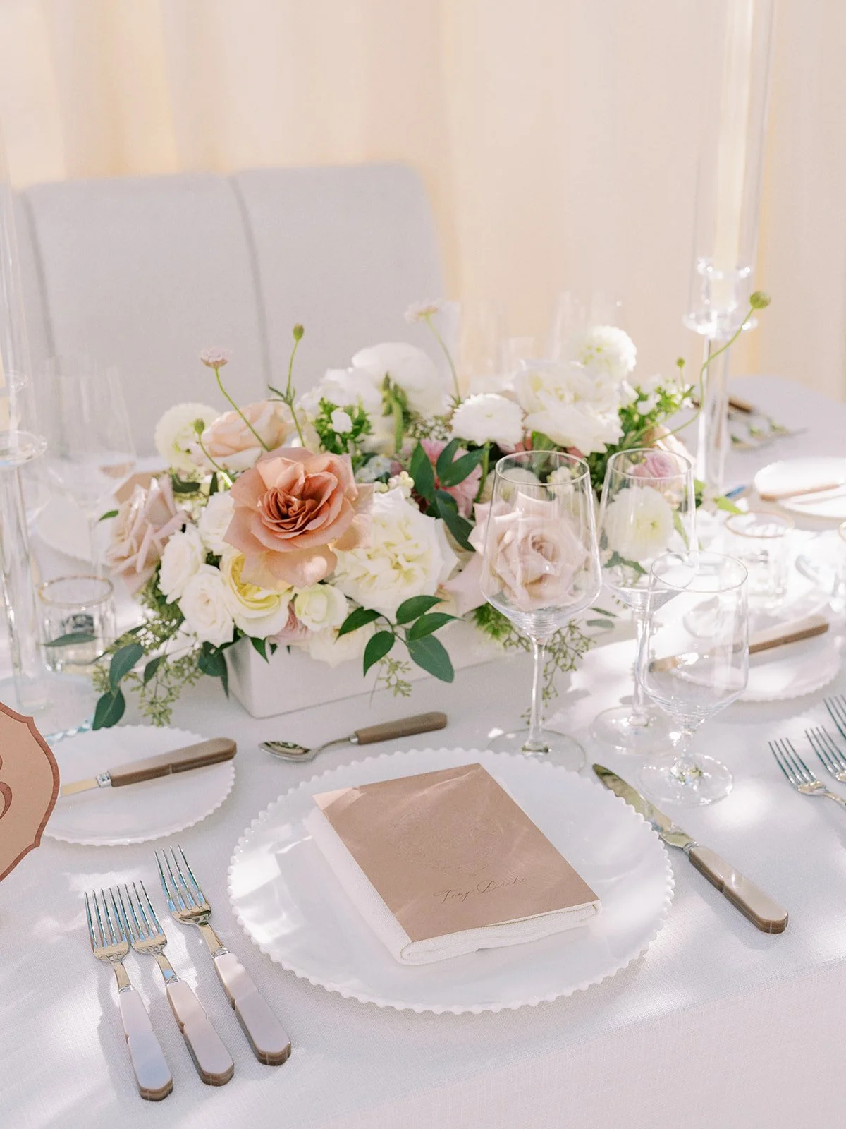 Elegant table setting with white tablecloth, plates, cutlery, and wine glasses. Centerpiece includes pink and white flowers. Tan-colored folded menu on the plate at Calgary tent wedding