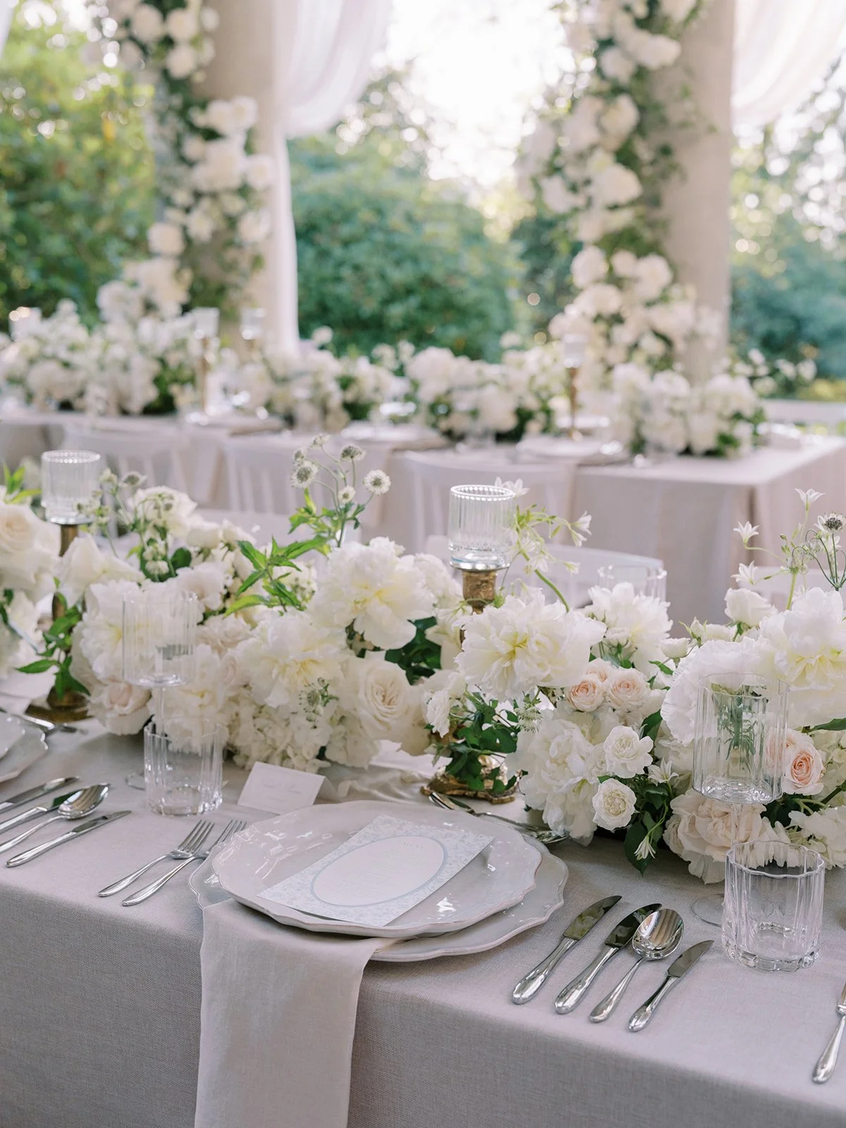 Elegant outdoor wedding reception table decorated with white flowers, candles, and fine tableware, under a canopy with white drapery and surrounded by lush greenery.