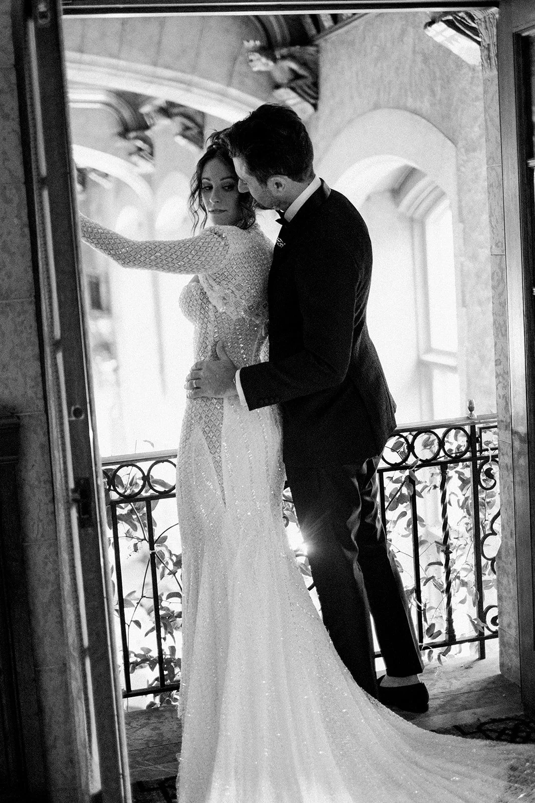 A couple in formal attire embracing on a balcony, with the woman wearing a beaded gown and the man in a suit. The setting is elegant and ornate.