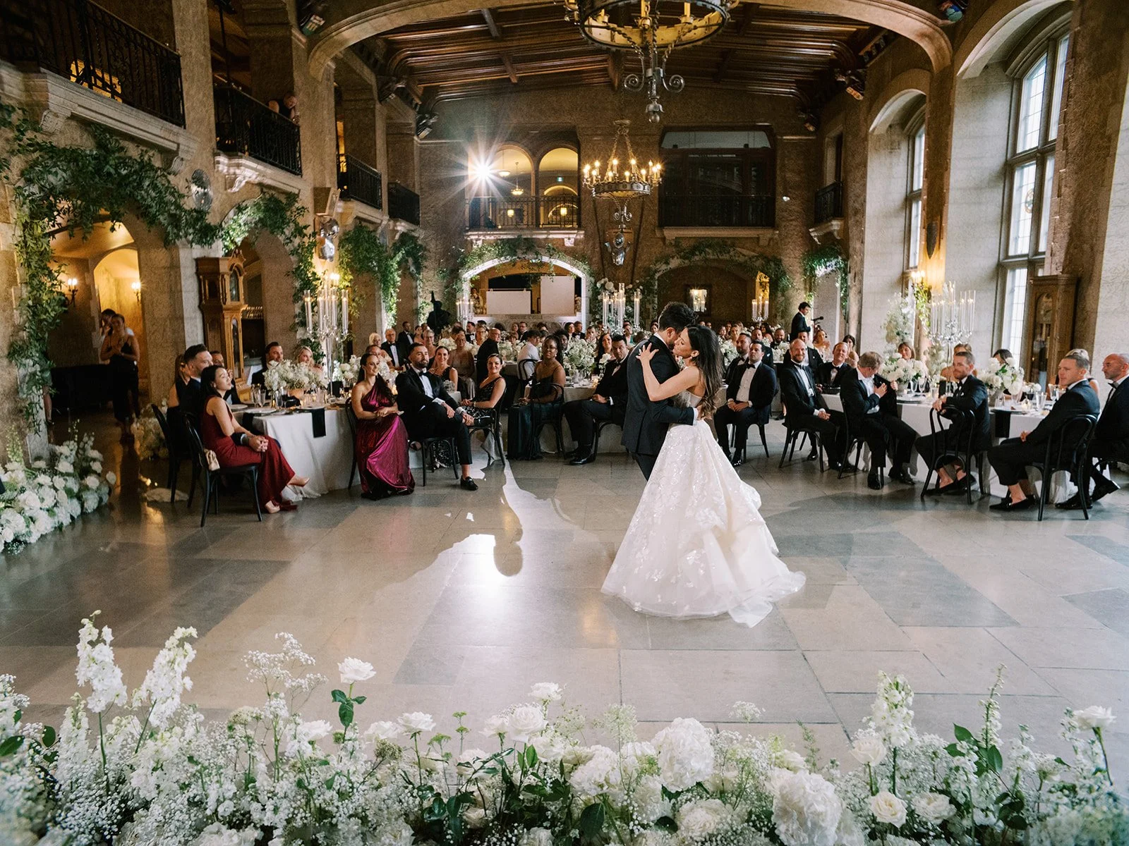 Bride and groom dancing at a wedding reception in an elegant venue with guests seated at decorated tables observing. The room features high ceilings, chandeliers, and floral arrangements in Mt.Stephen Hall at The Fairmont Banff Springs