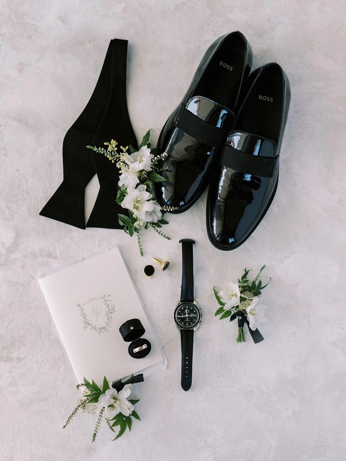 Elegant black men's shoes, black bow tie, wristwatch, cufflinks, wedding ring, boutonniere, and a white floral-themed invitation card on a textured surface.