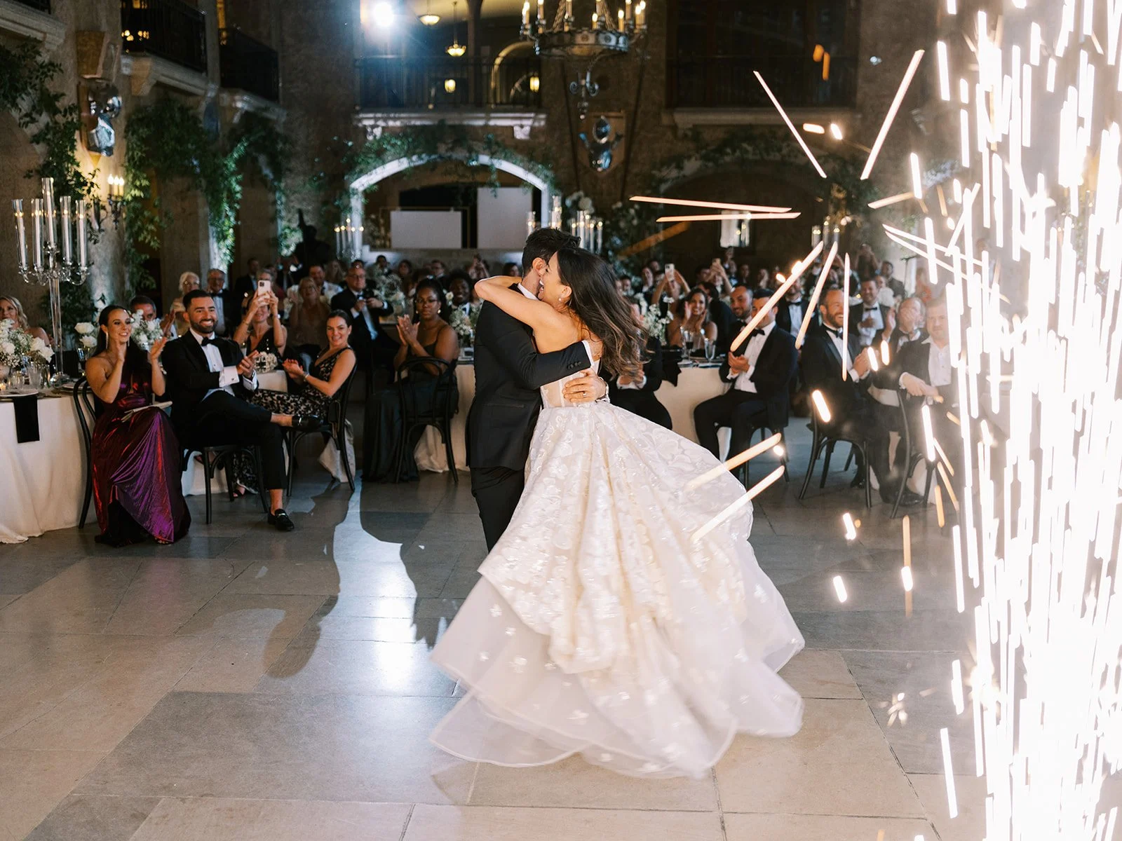 A couple dancing at their wedding reception at the Banff Springs Hotel with guests applauding. The bride wears a white gown and the groom a black suit. Indoor venue with a festive atmosphere and sparklers in the foreground.