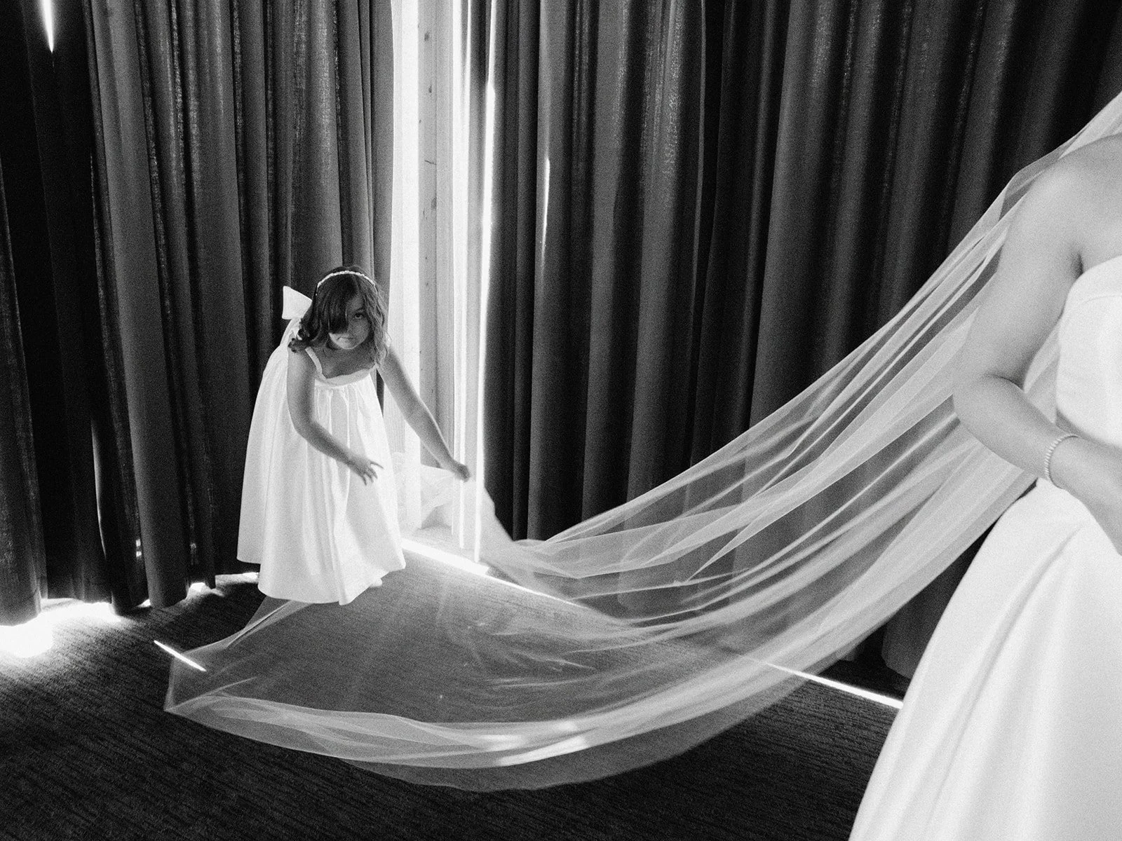 Black and white photo of a flower girl holding a bride's veil, with curtains in the background.