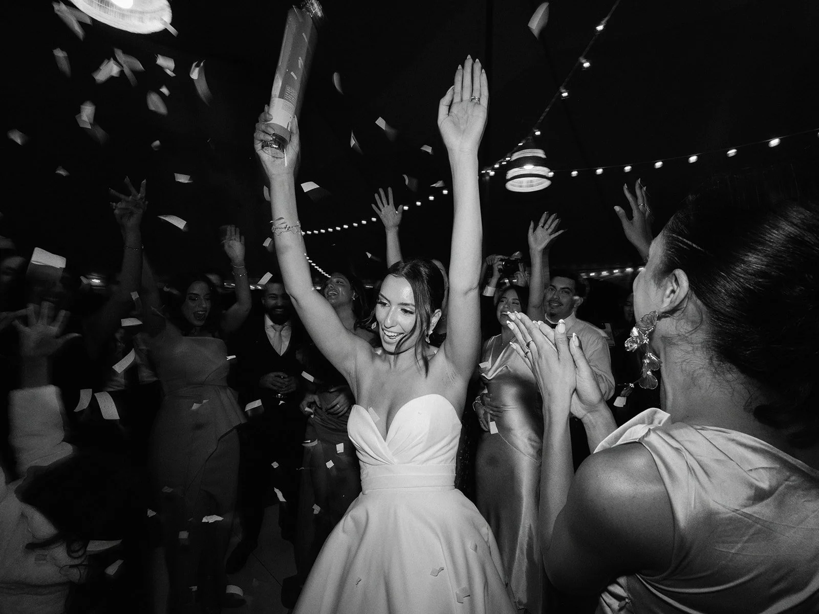A joyful woman in a strapless dress raising her arms, surrounded by a group of people celebrating with confetti falling, under string lights in a festive atmosphere at a tent wedding in Calgary