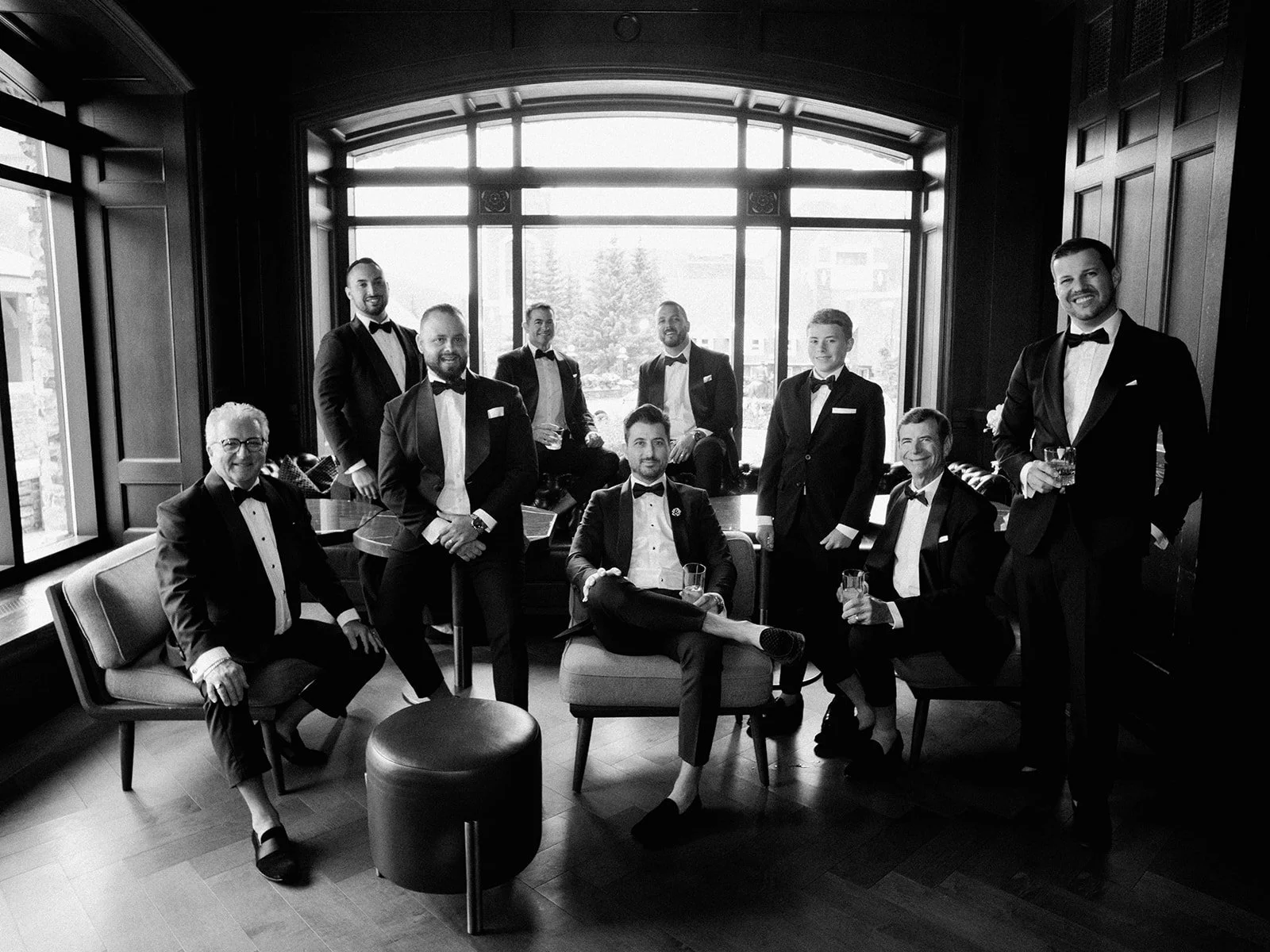 Group of men in tuxedos posing in a room with large windows and wood paneling.