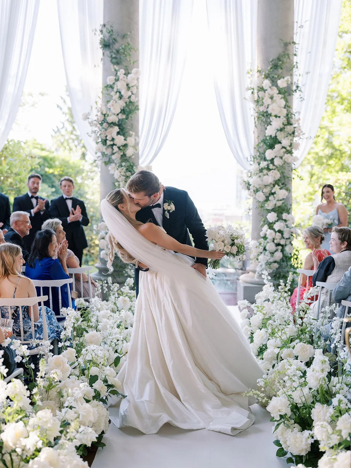 A newlywed couple sharing a kiss during their wedding ceremony, surrounded by white flowers, with guests clapping and watching.