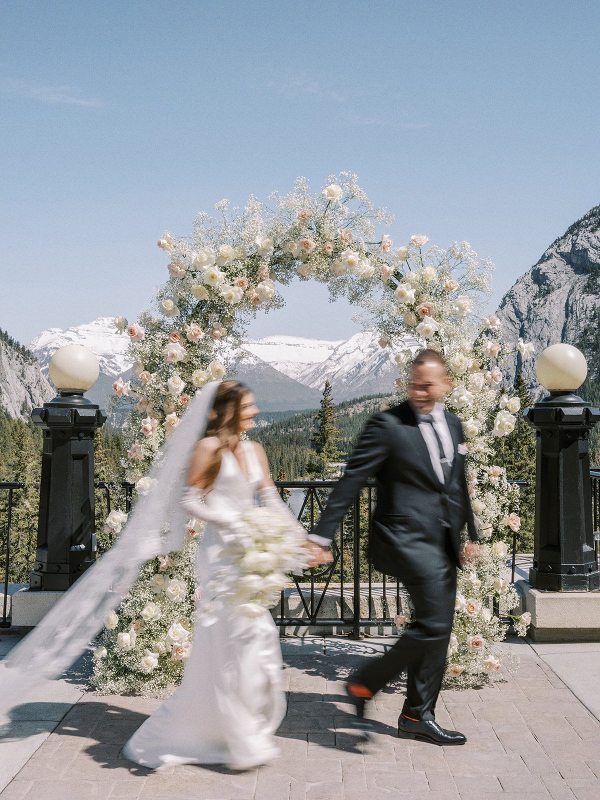 A couple in wedding attire walking hand in hand past a floral arch with mountains in the background under a clear sky.