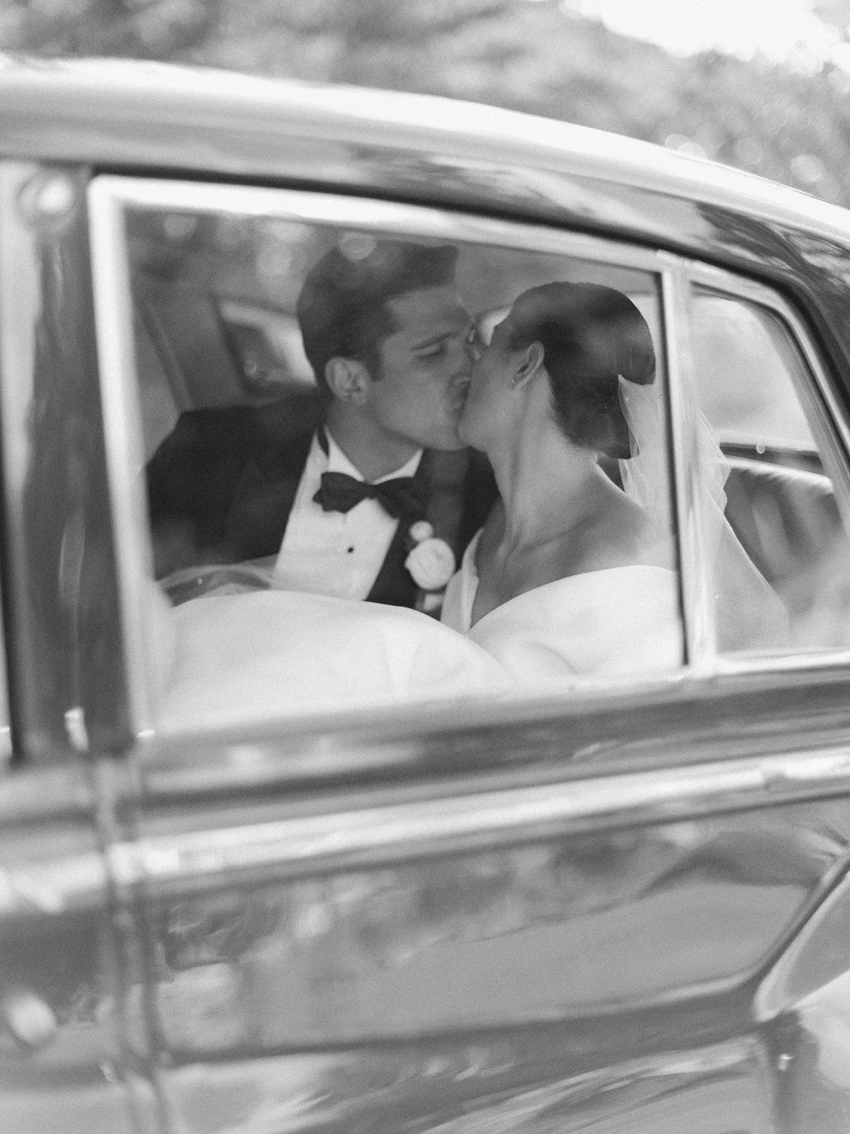 Bride and groom kissing in a car, photographed through the window, in black and white.