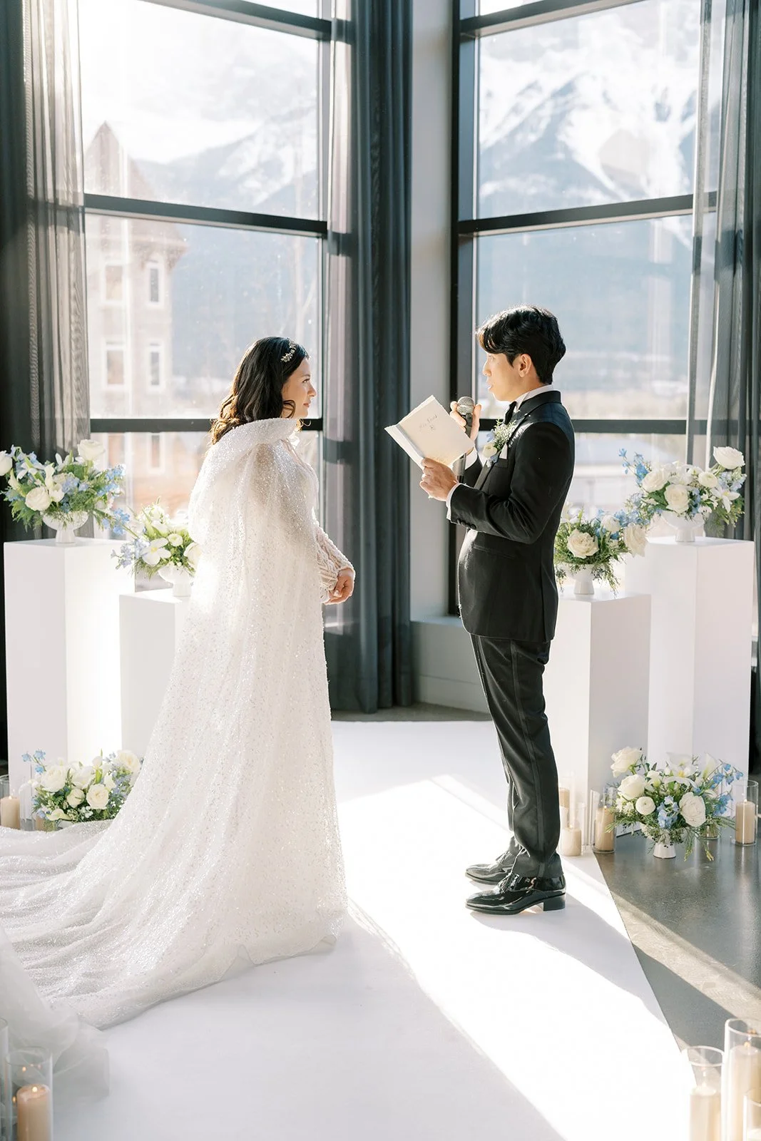 Bride and groom exchanging vows indoors with floral decorations and mountains visible through large windows at Mainspace by Canmore wedding photographers