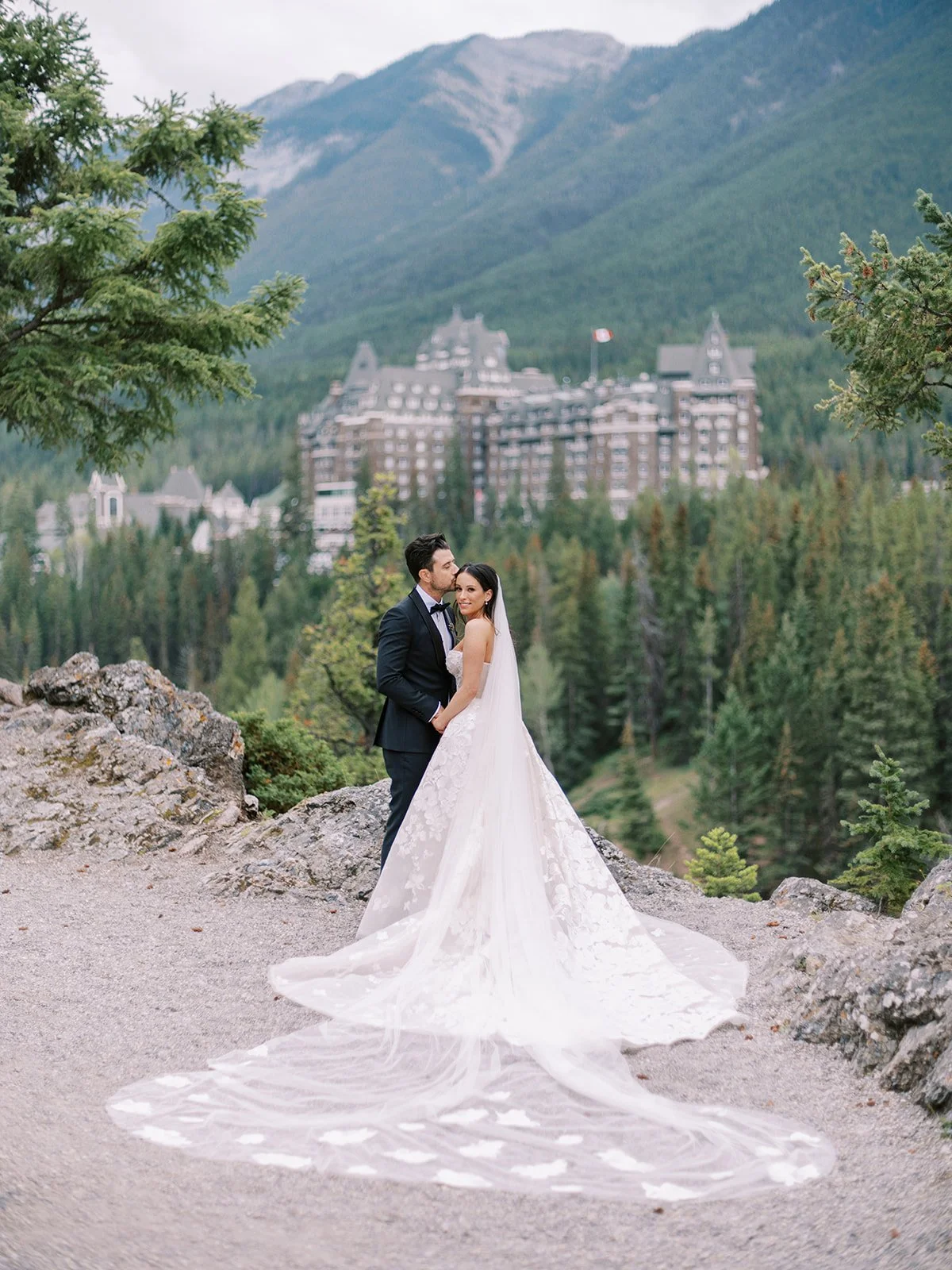 Bride and groom embracing outdoors with a forest and mountain in the background, wearing wedding attire. At Surprise Corner in Banff overlooking the Banff Springs Hotel.
