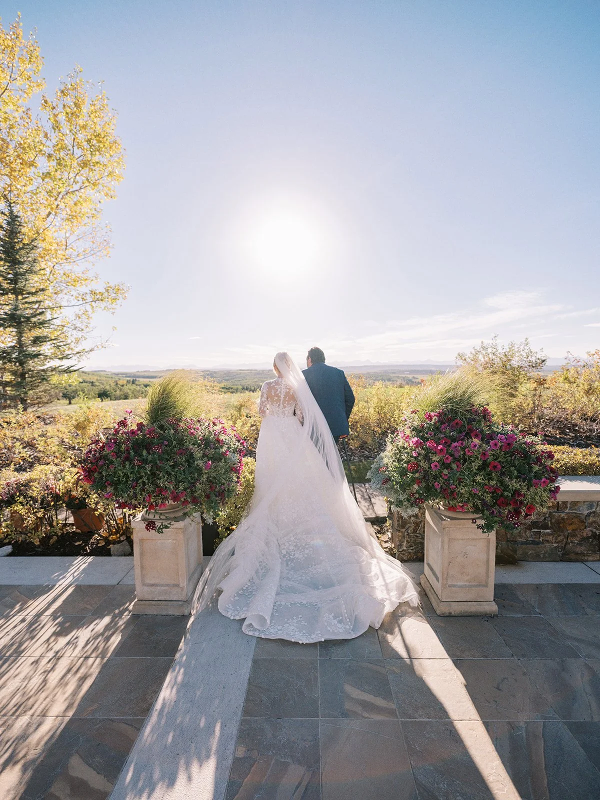 A bride and groom standing together outdoors at sunset, facing away, with flower arrangements on either side.