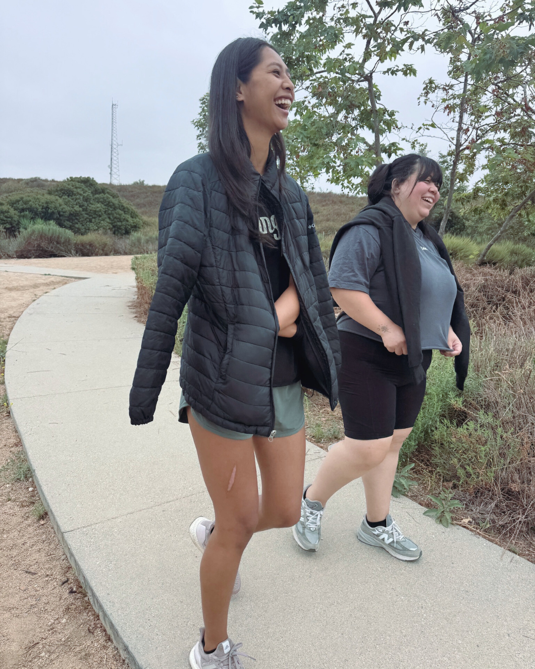 Two smiling women walking on a curved sidewalk in a park, surrounded by greenery, wearing casual outdoor clothing.