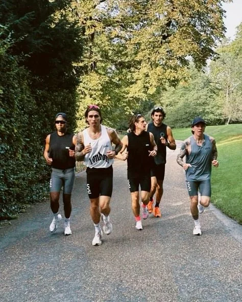 Five young adults running together on a paved park path surrounded by trees and greenery.