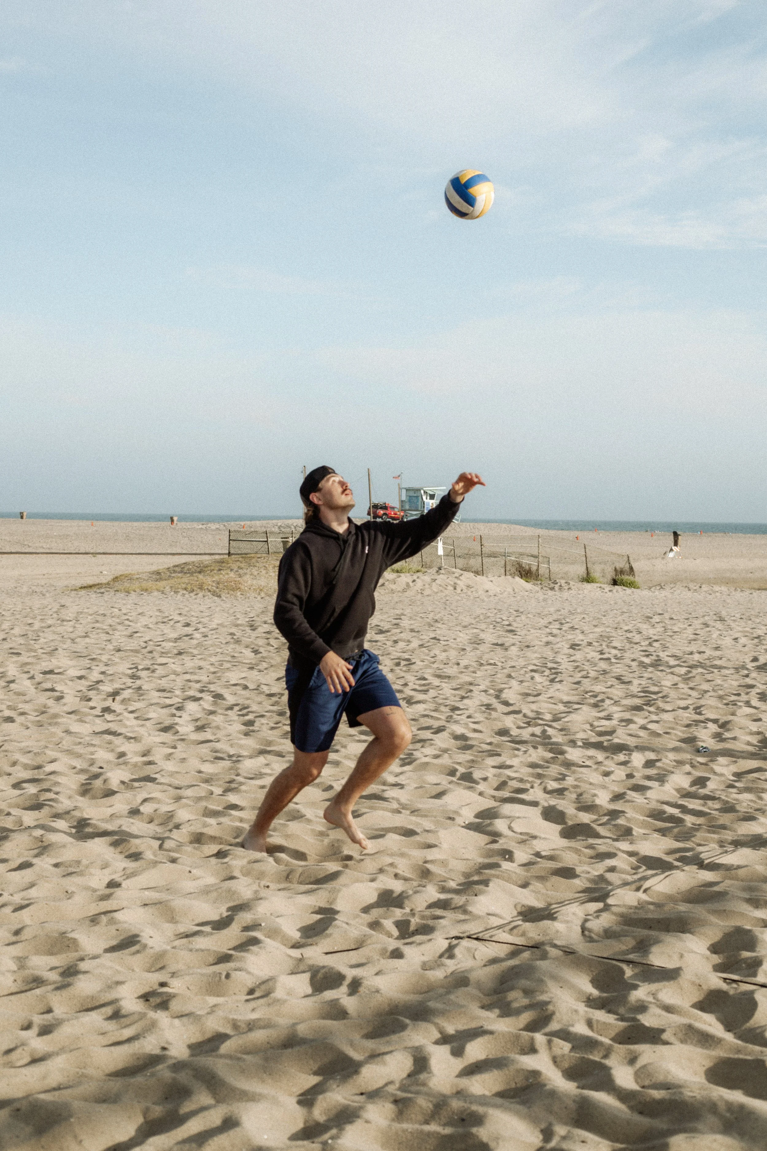 A young man playing beach volleyball on the sand, with the ocean and a lifeguard tower in the background.